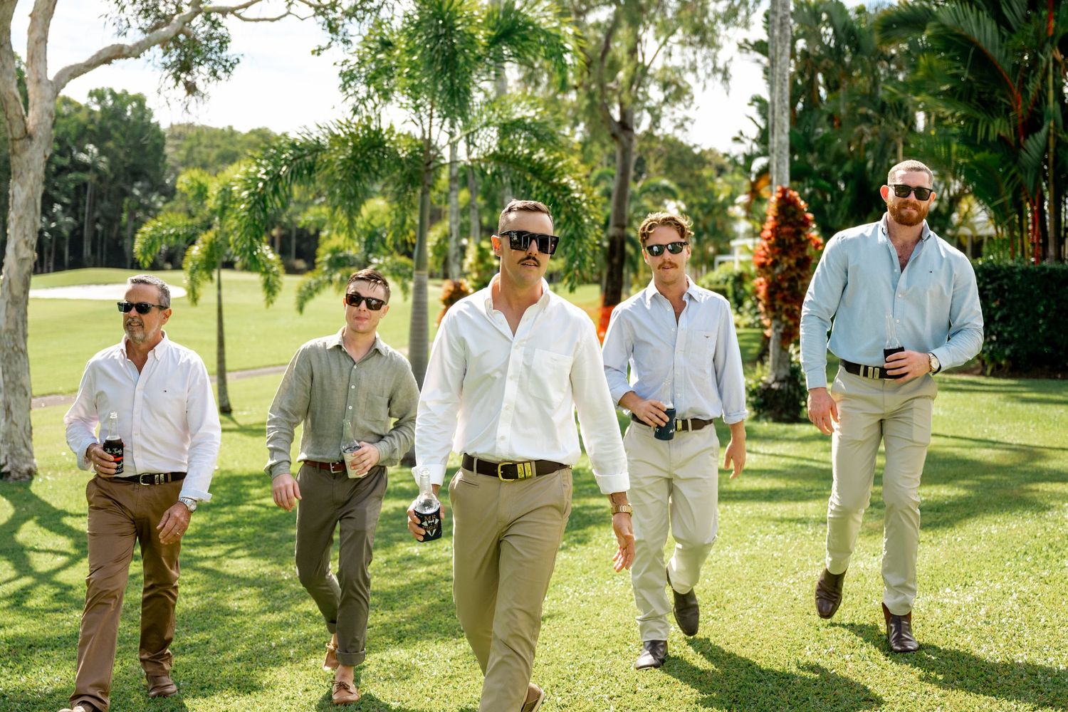 Five groomsmen in white shirts and khaki pants walking confidently across green lawn at outdoor wedding.