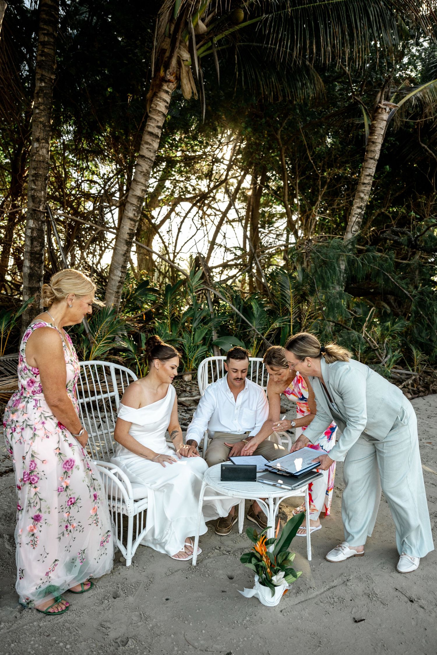 Wedding party gathered around bride and groom signing marriage documents on a tropical beach at Four Mile Beach, Port Douglas.