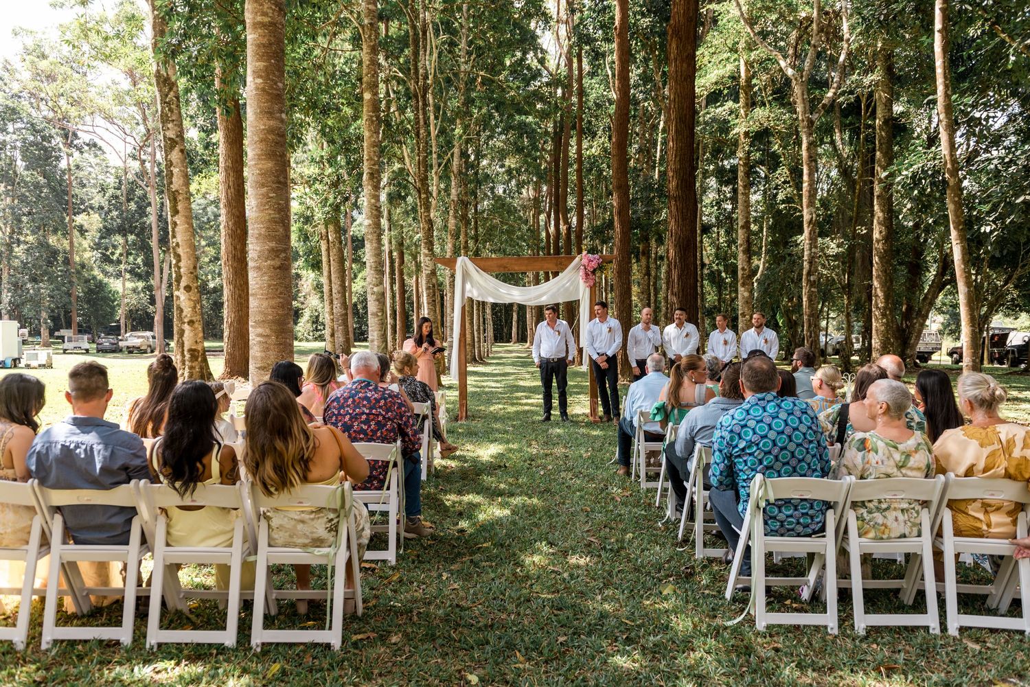 Outdoor wedding ceremony in a tree-lined grove with white chairs and groomsmen at the altar.