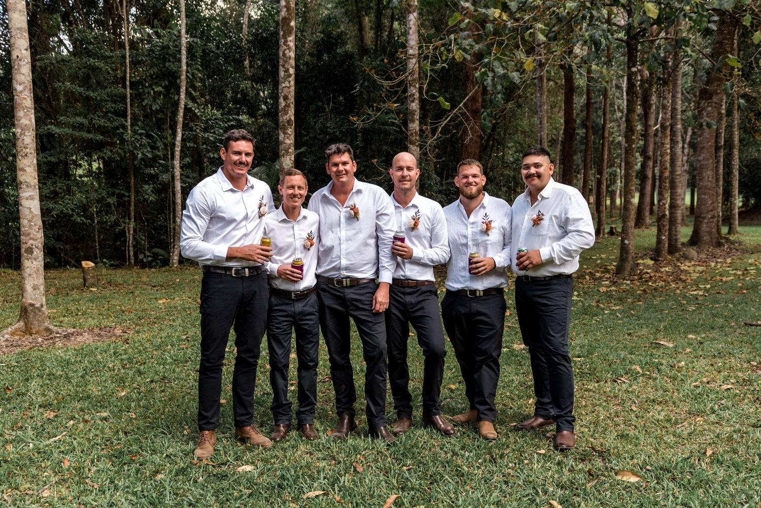 Six groomsmen in white shirts and dark pants toasting with drinks at outdoor forest wedding.
