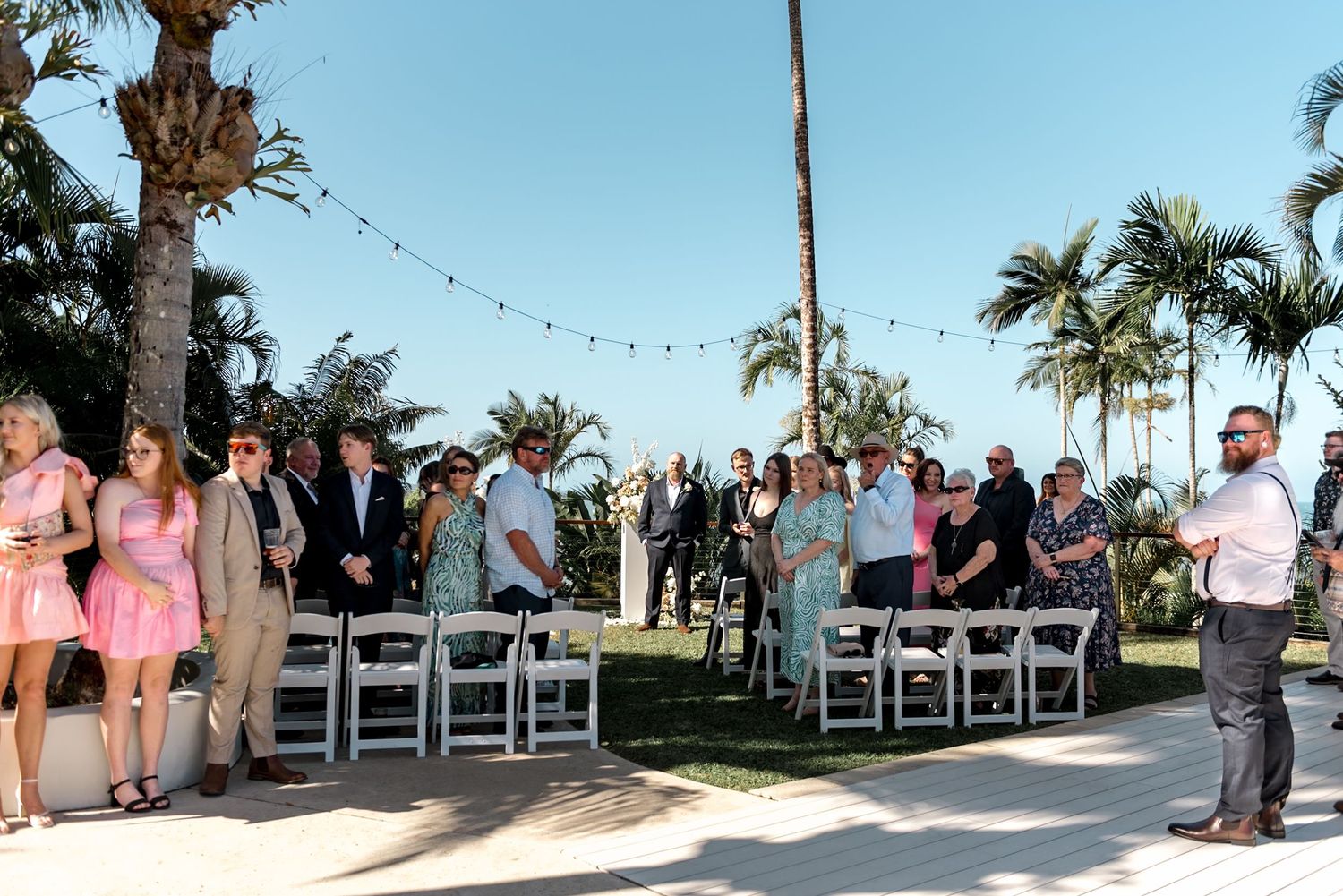 Wedding guests stand for outdoor tropical ceremony with white chairs, palm trees, and string lights.