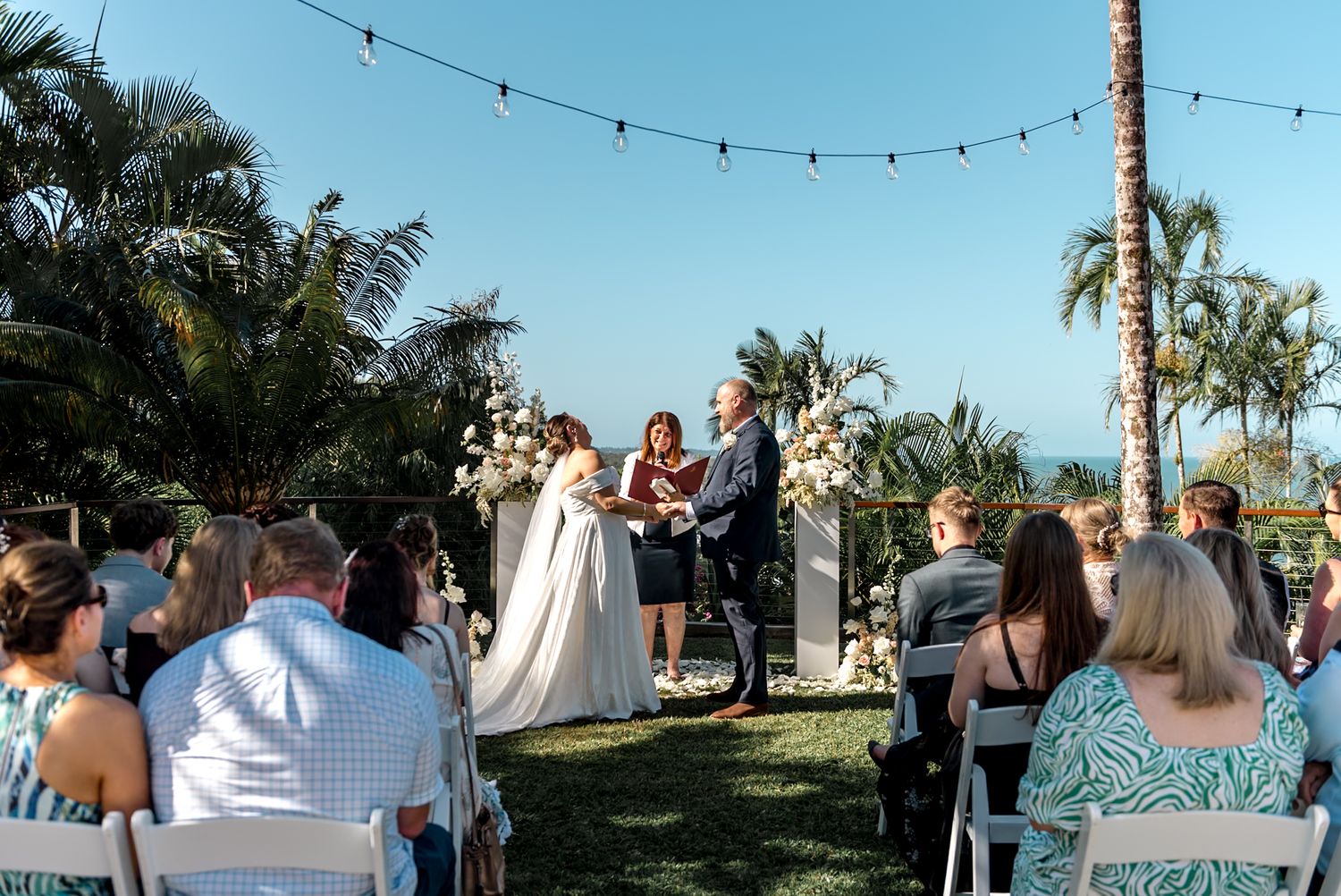 Guests seated in white chairs watch bride and groom exchange vows at tropical outdoor wedding ceremony.