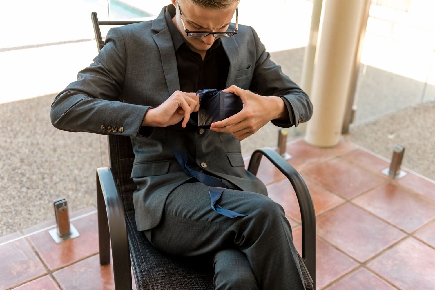 Man in gray suit adjusting his tie while seated in a chair indoors.