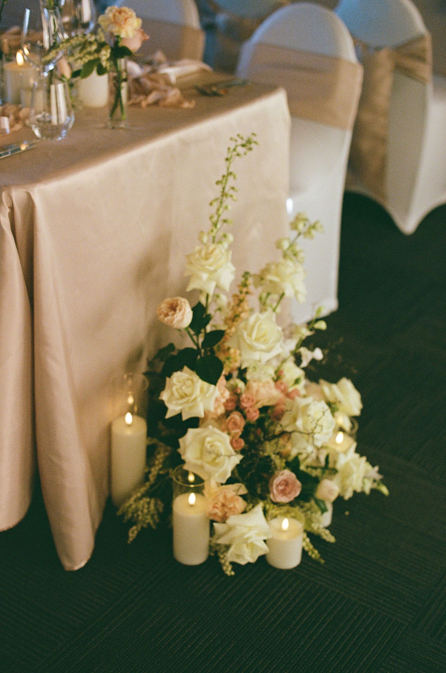 Elegant wedding floor arrangement of white and blush roses with pillar candles beside a reception table.