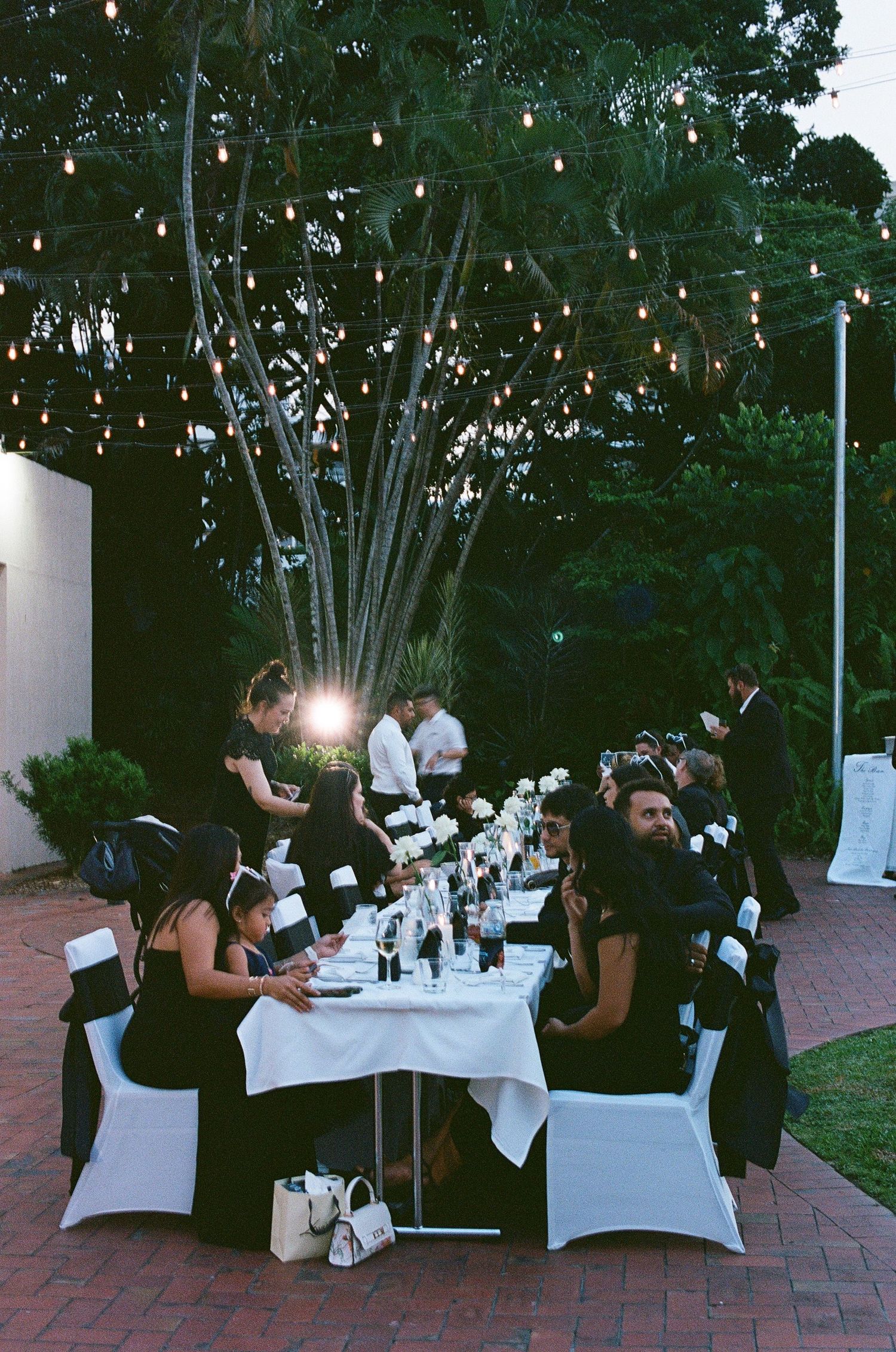 Guests seated at outdoor wedding reception under warm string lights at dusk.