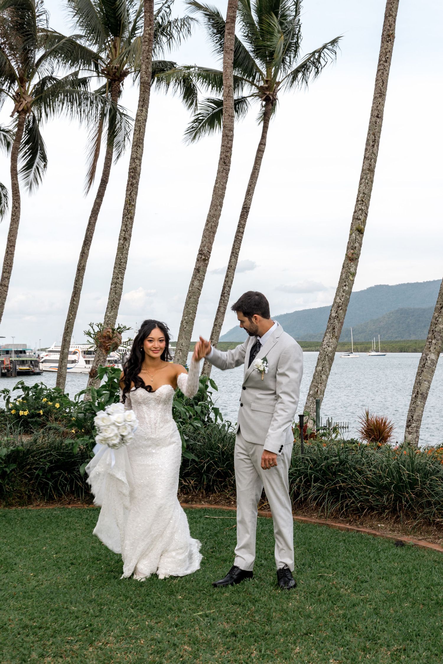Groom twirls bride in strapless lace gown near waterfront with tall palm trees in background.