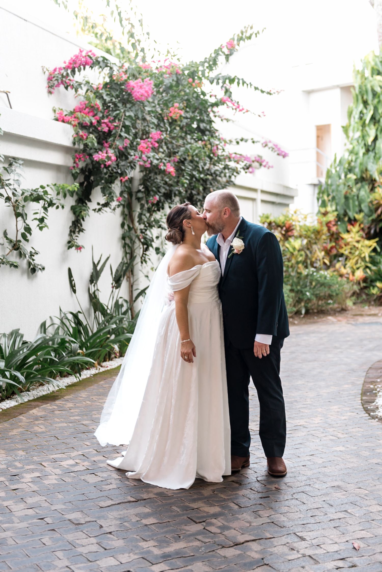 Bride and groom sharing a kiss on brick courtyard with pink bougainvillea climbing white wall.