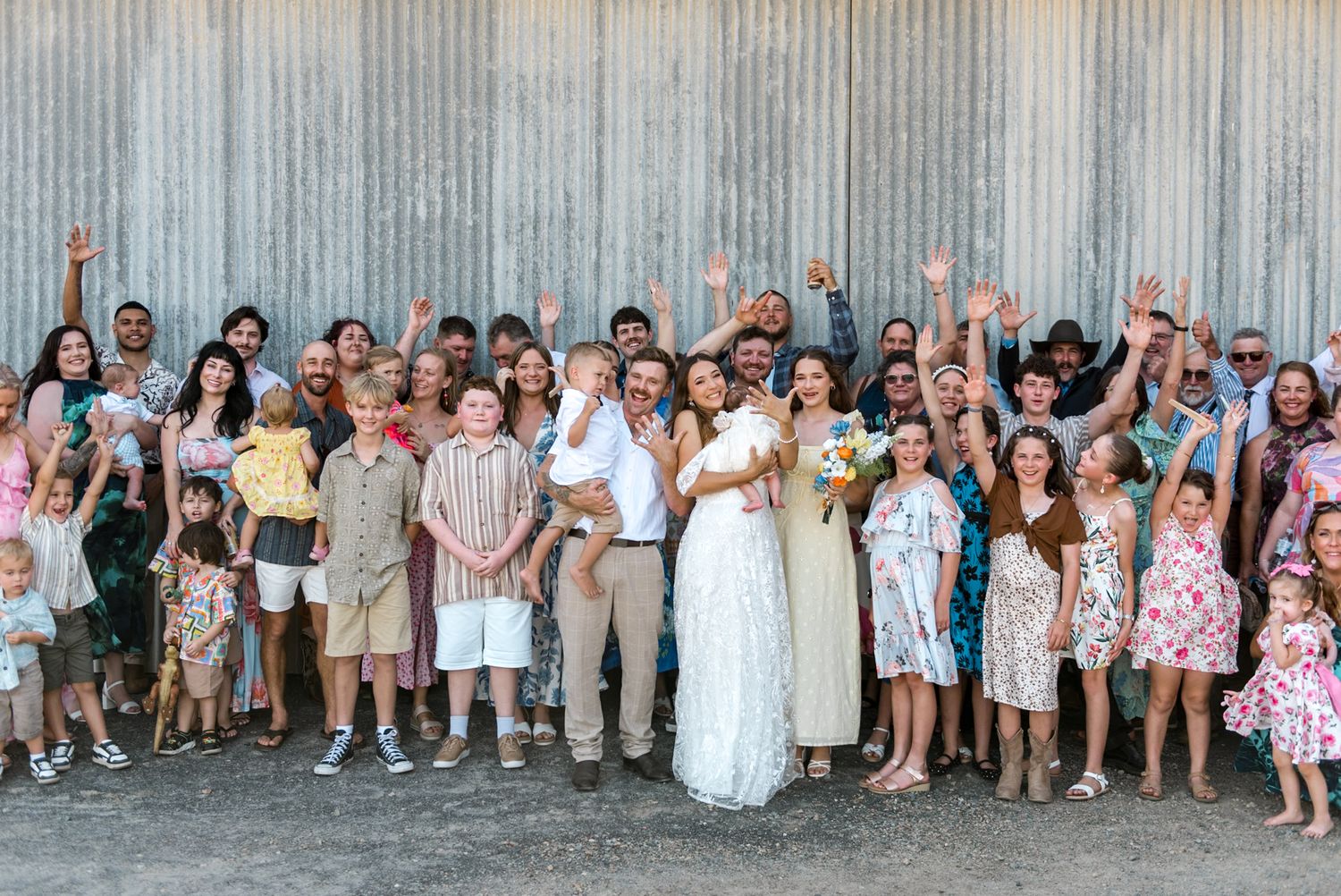 Joyful wedding group photo with bride, groom and guests cheering with raised arms outside a barn.