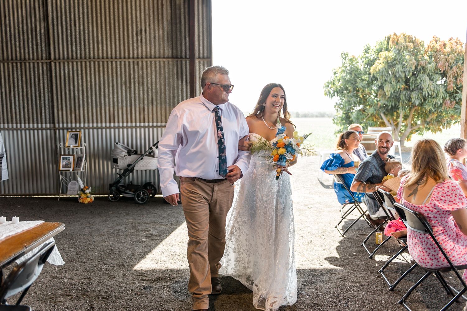 Bride in lace gown walks down aisle with escort inside a rustic barn wedding venue.