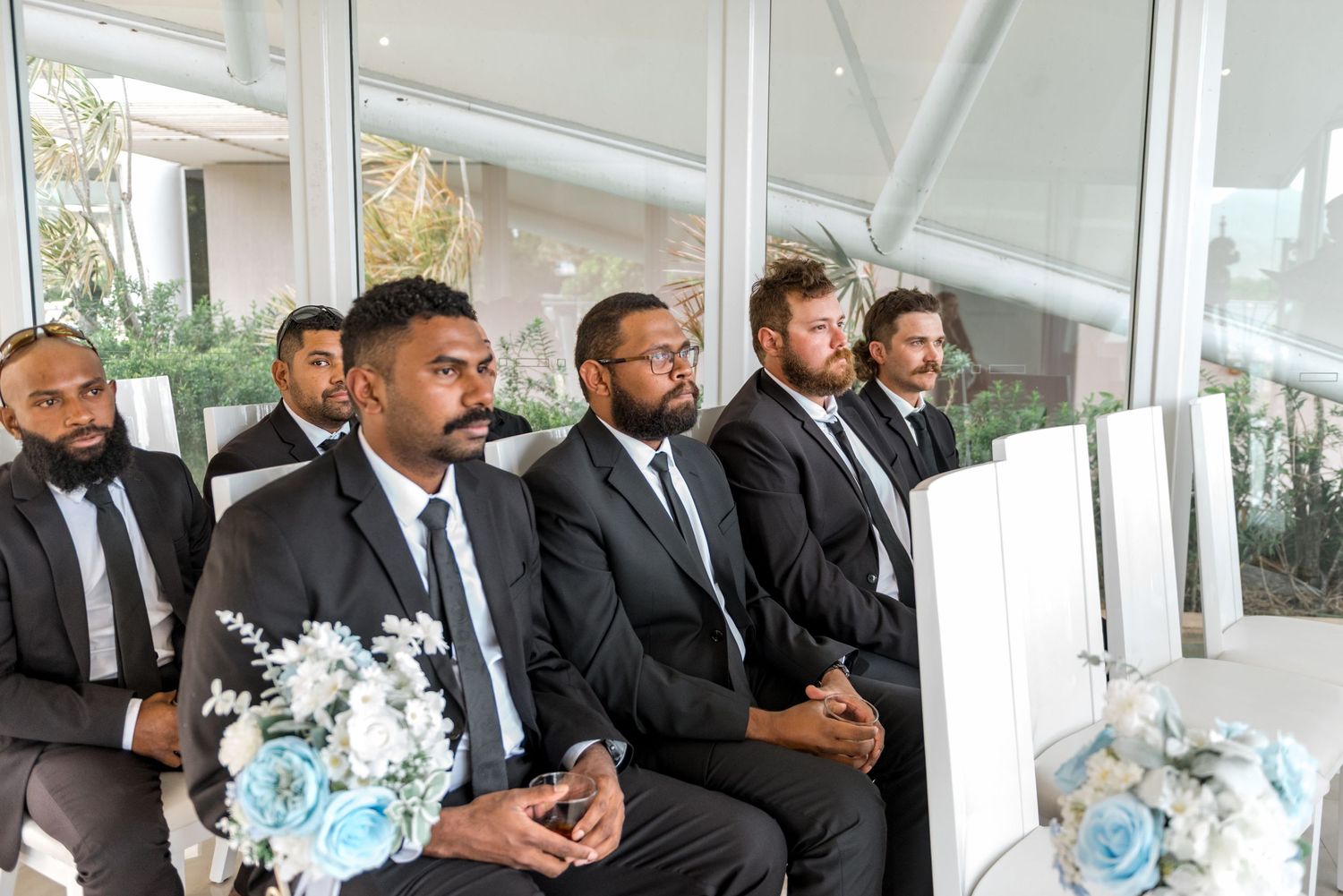 Groomsmen in black suits seated at a wedding ceremony with blue floral arrangements.