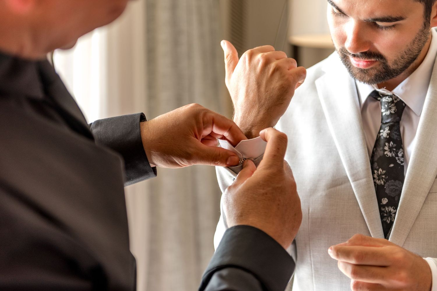 Groom smiling as someone helps fasten his cufflinks while dressed in a light gray suit and floral tie.
