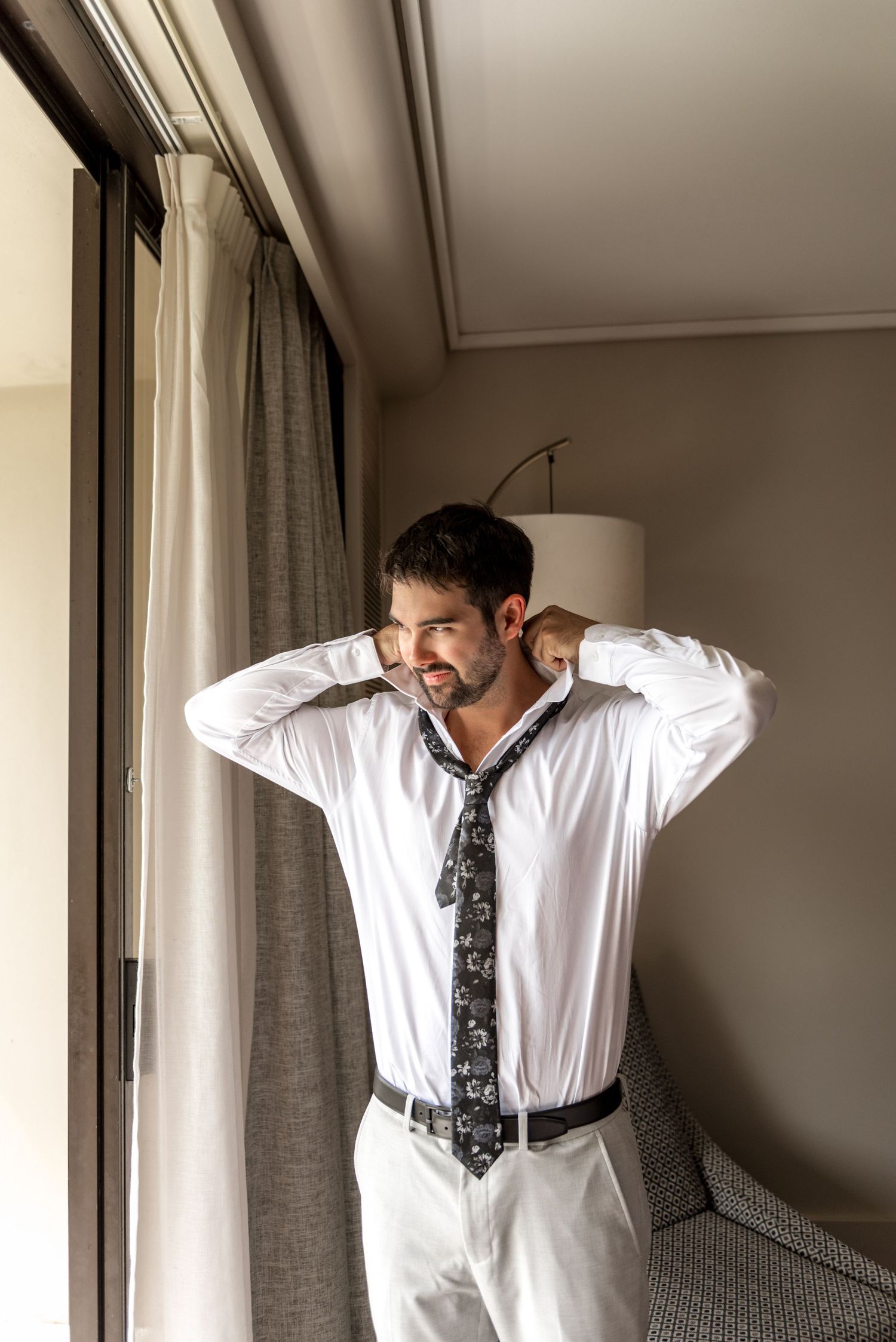 Groom in white shirt and patterned tie posing by hotel window before wedding.