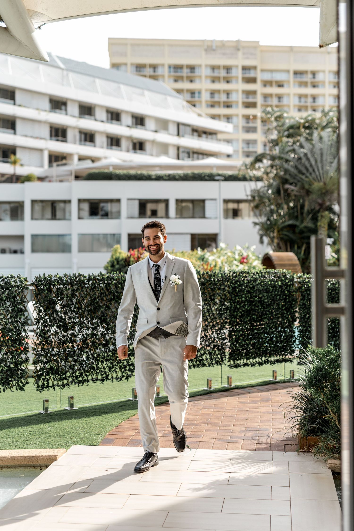 Groom in light grey suit walks through outdoor hotel courtyard on wedding day.