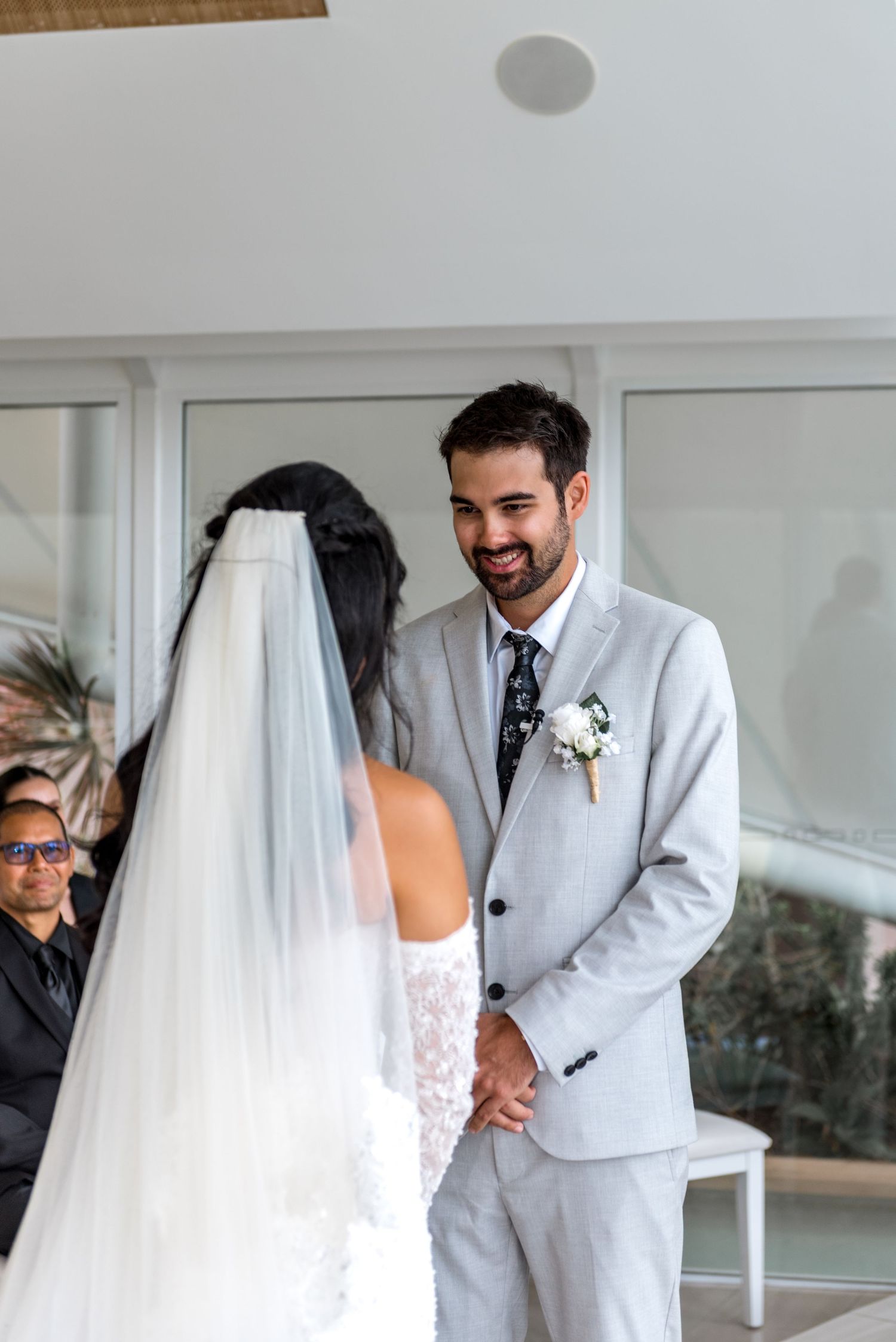 Groom in gray suit smiling joyfully at bride wearing white veil during wedding ceremony.