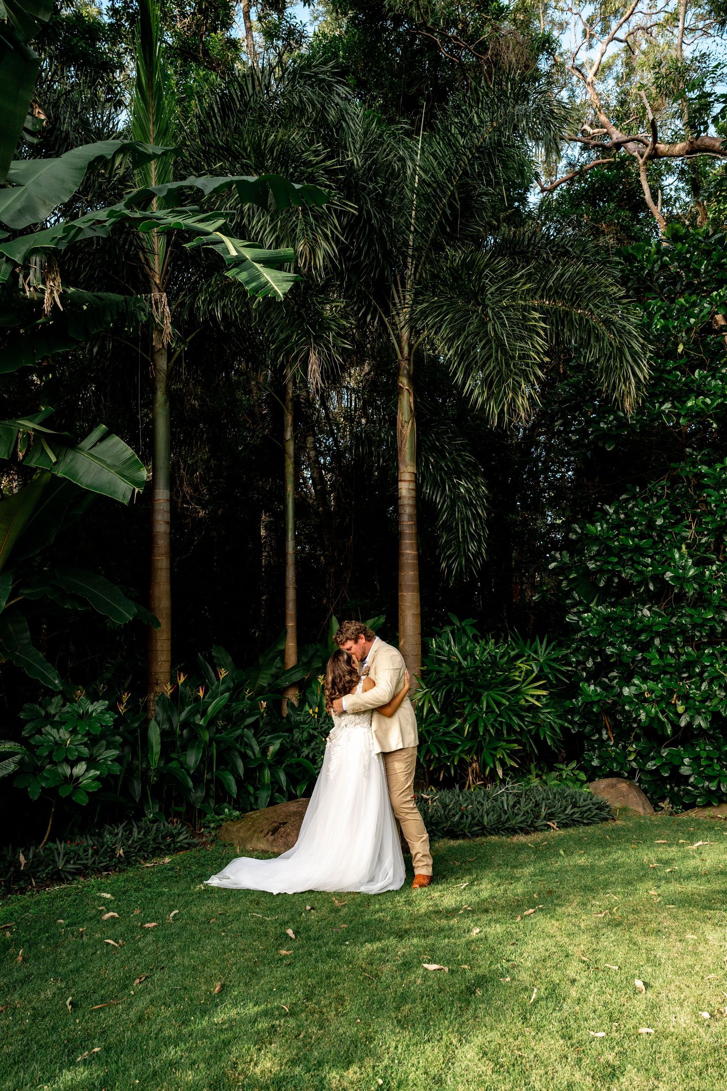 Bride and groom share tender kiss surrounded by tall palm trees in tropical garden setting.
