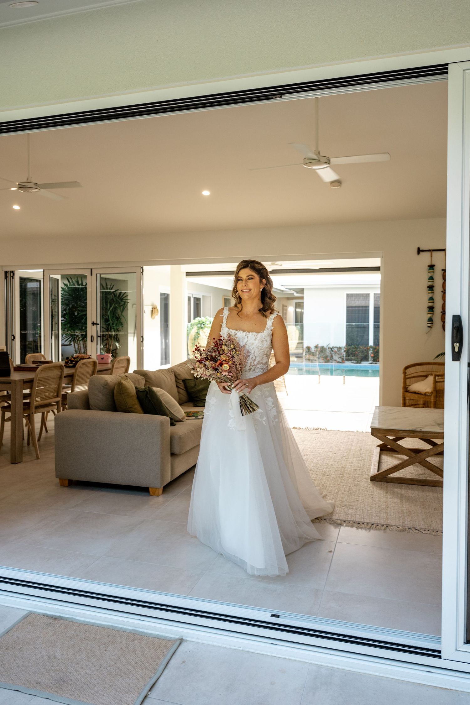 A bride in a white tulle gown holds a bouquet while standing in an open sliding glass doorway.