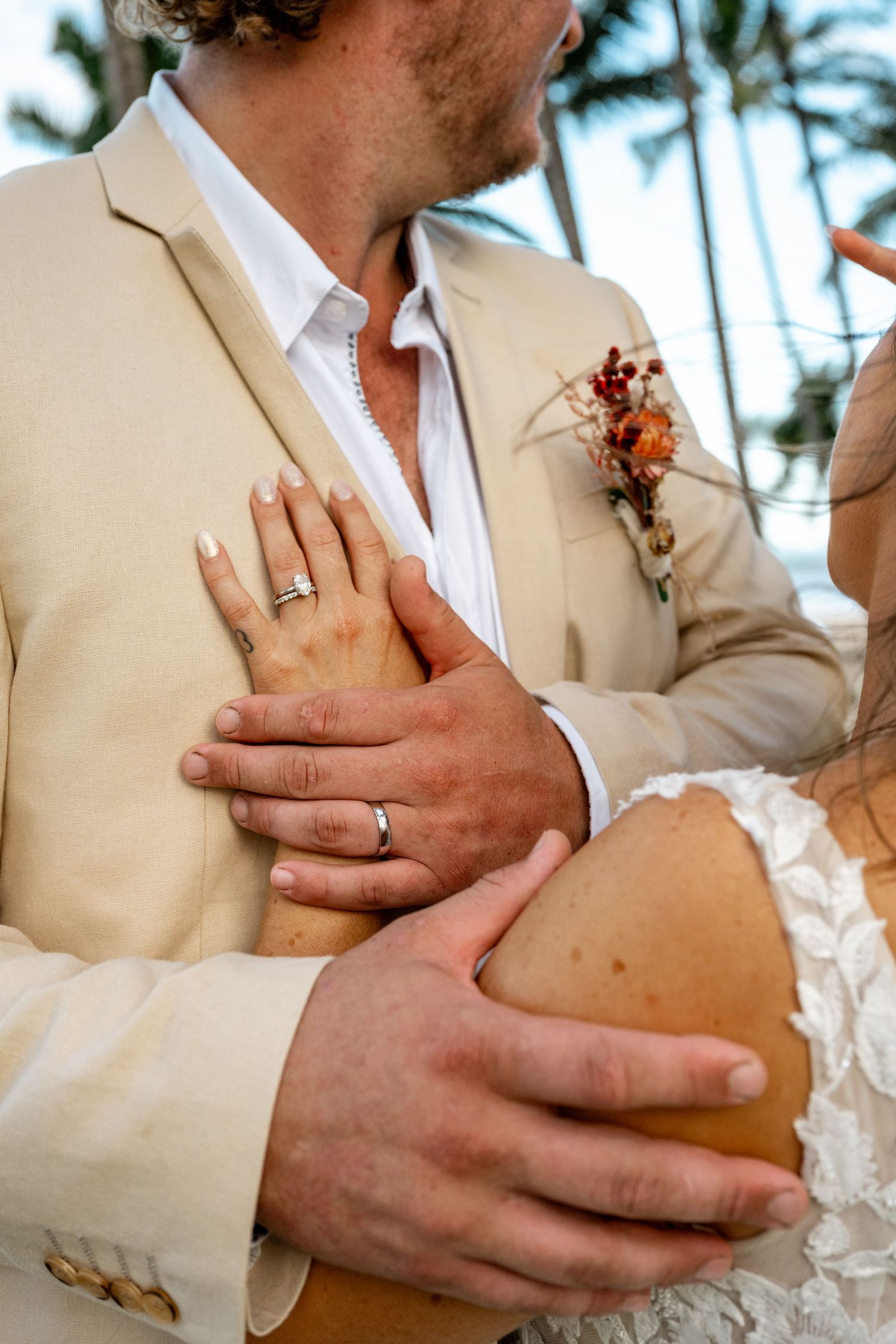 Close-up of bride and groom embracing, showcasing wedding rings and groom's floral boutonniere in tropical setting.