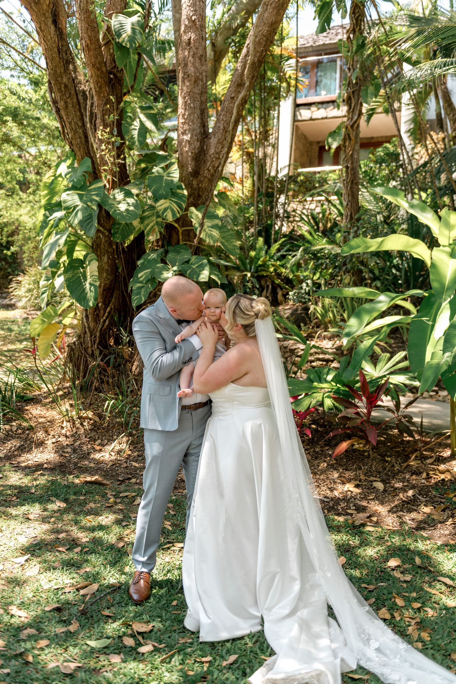 Bride and groom in tropical garden tenderly holding their baby on their wedding day.