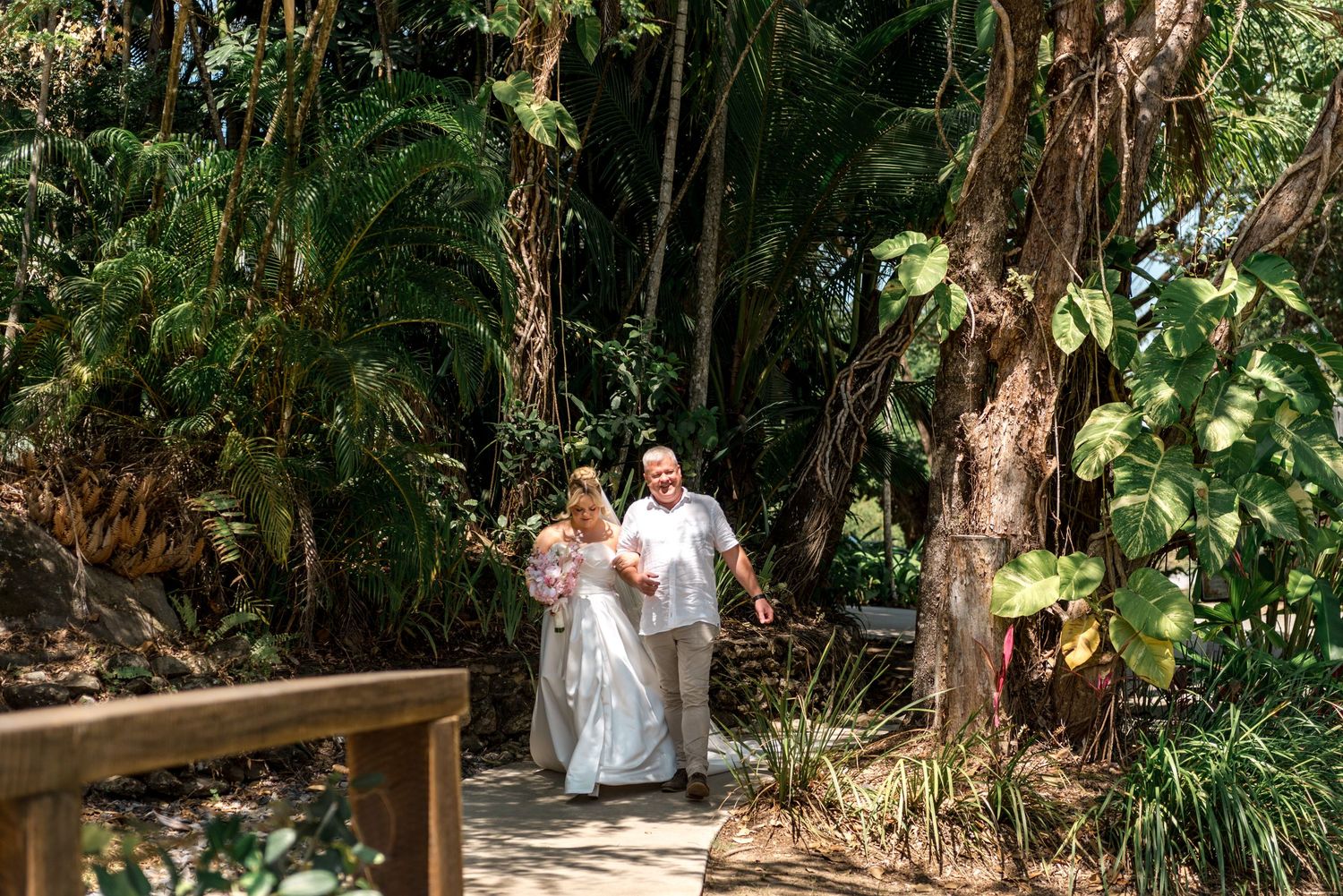 Bride in white gown and groom stroll through lush green tropical wedding garden setting.