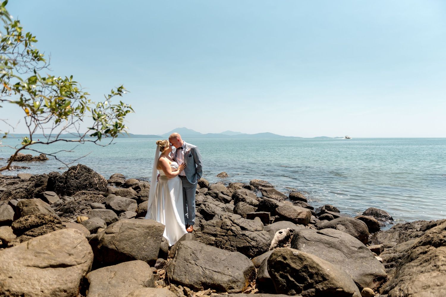 Bride and groom share a romantic kiss on rocky shoreline with turquoise ocean and mountains behind.