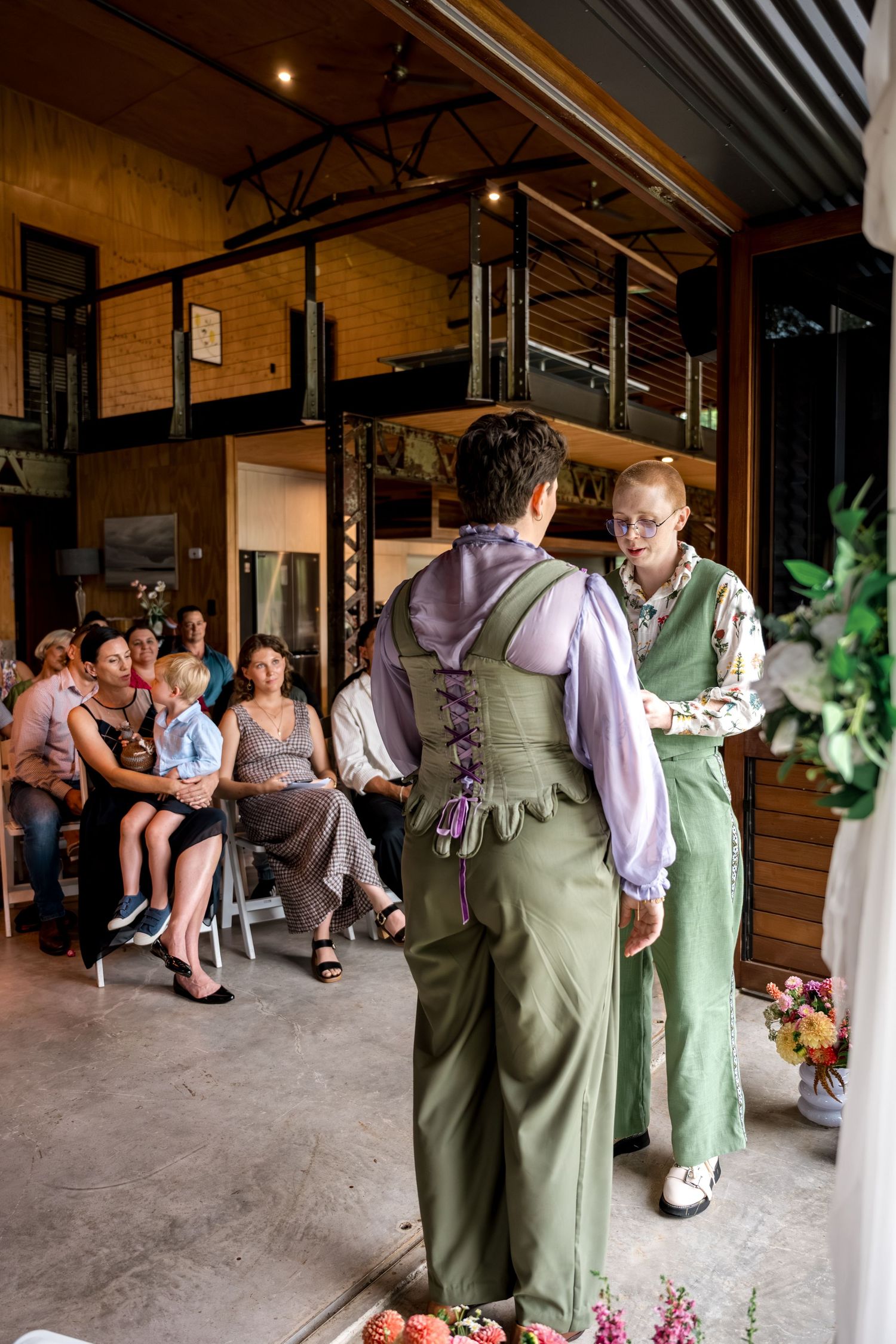 Two brides in green overalls and purple blouses exchange vows at an indoor wedding ceremony with seated guests.