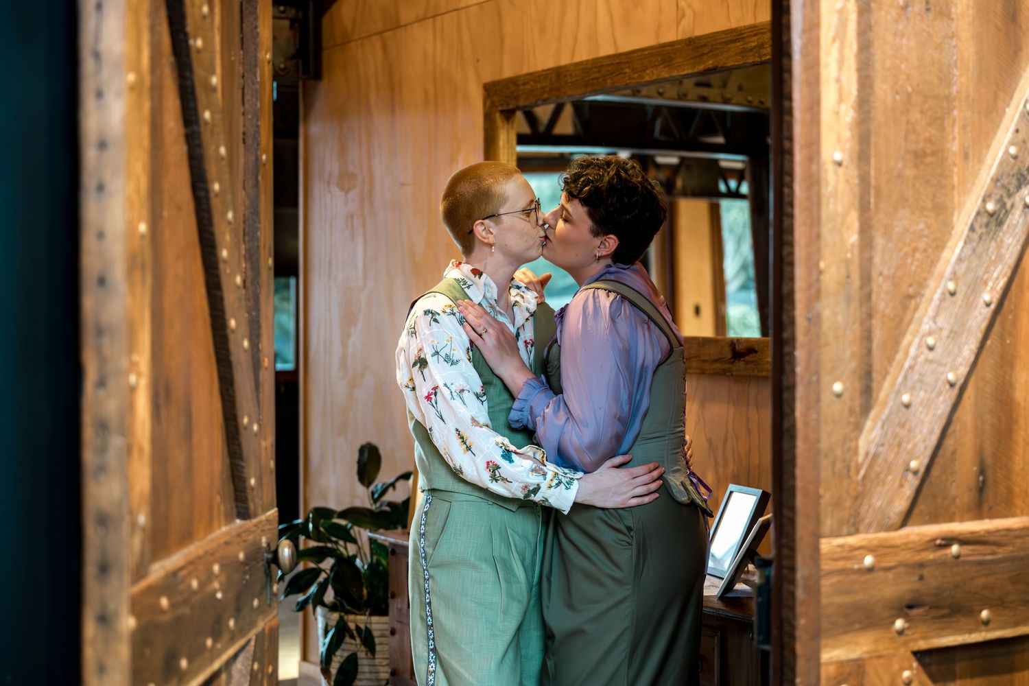 Two women share a kiss in a doorway of a warmly lit rustic wooden building.