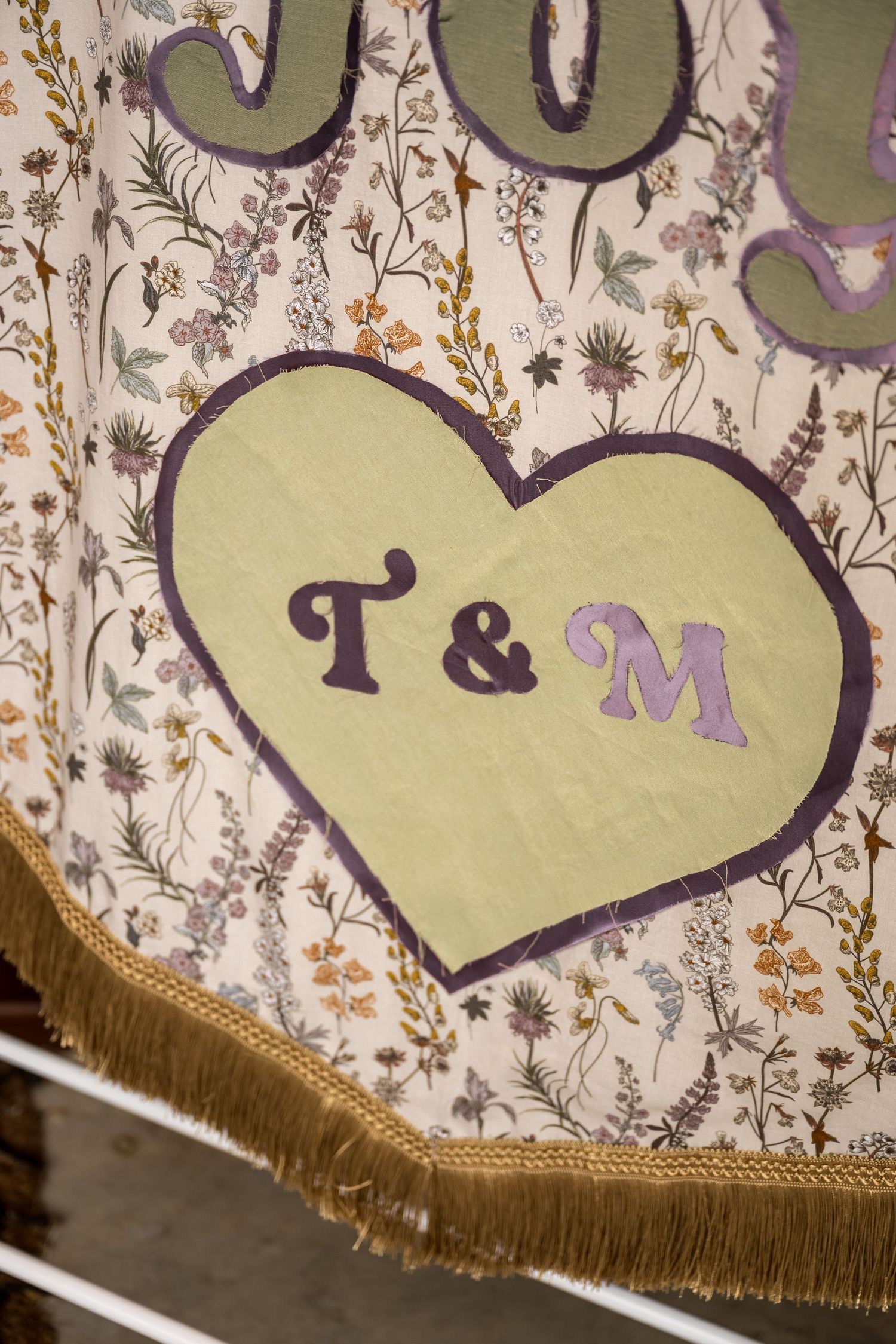 Floral fabric banner featuring a green heart with T&M initials and gold fringe trim.