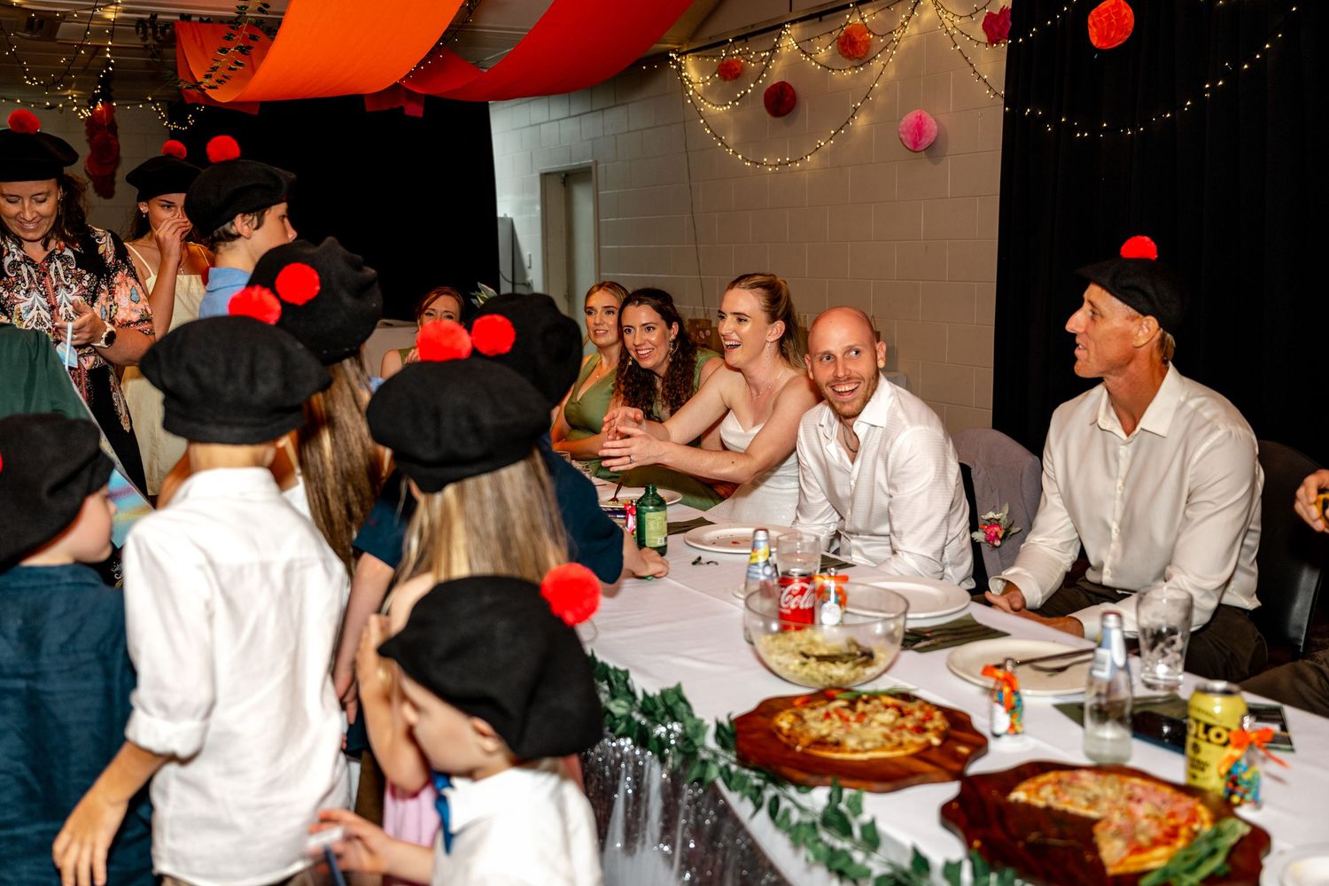 Bride and groom seated at wedding reception table surrounded by guests in festive costumes.