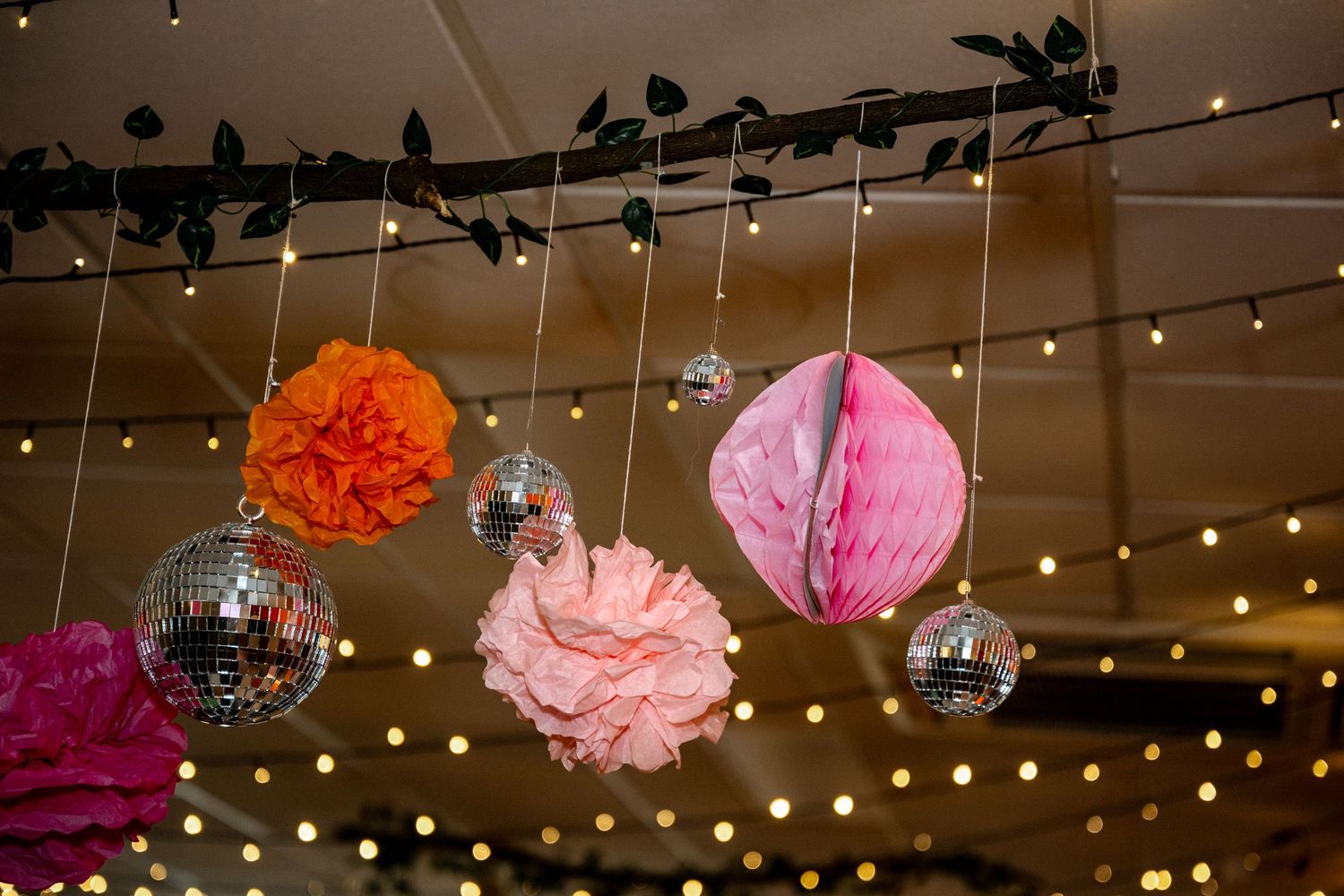 Disco balls and colorful tissue paper pom-poms hanging from ceiling with fairy lights at wedding reception venue.