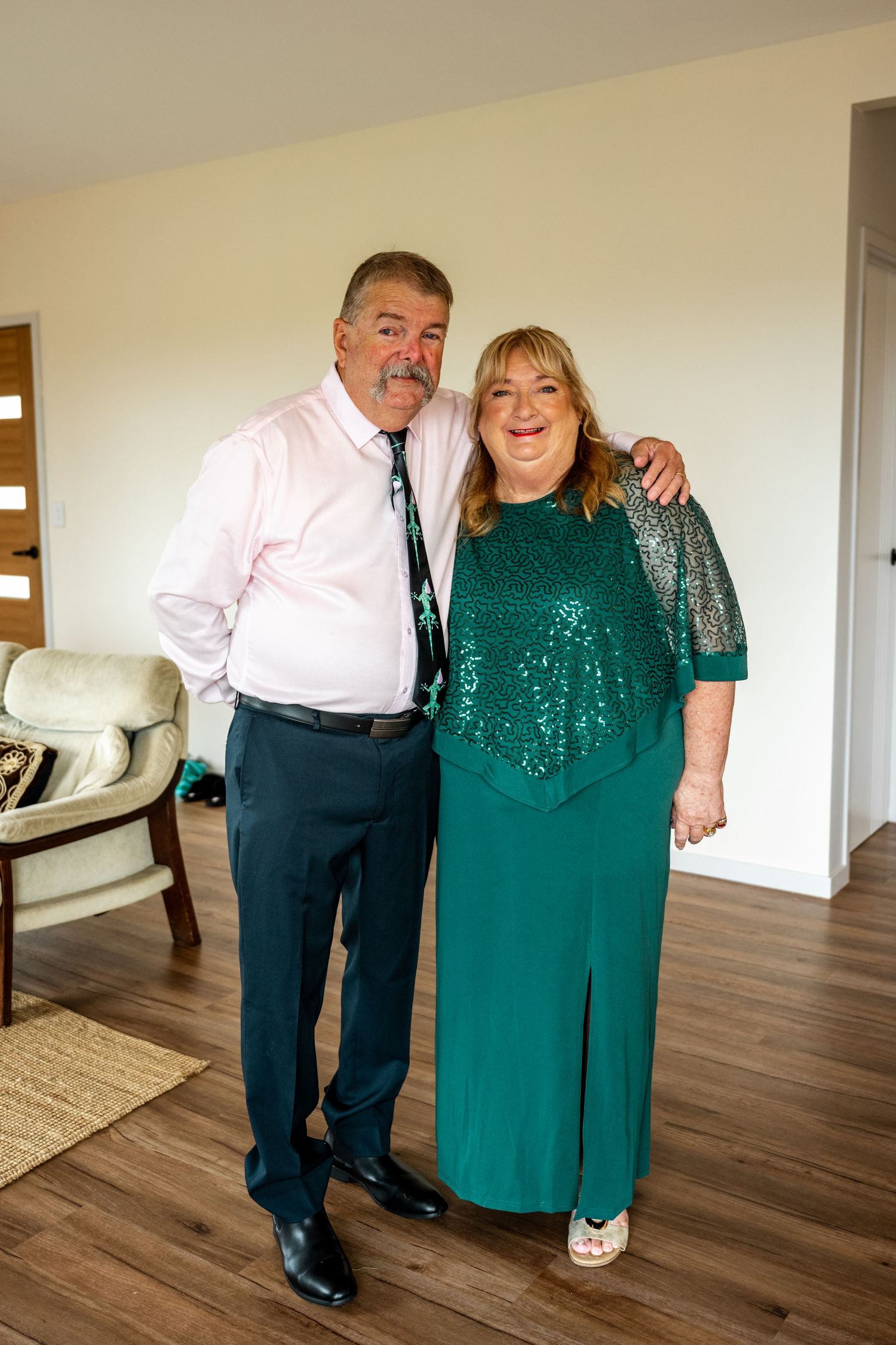 Smiling couple dressed in green sequin gown and pink shirt posing together in living room.