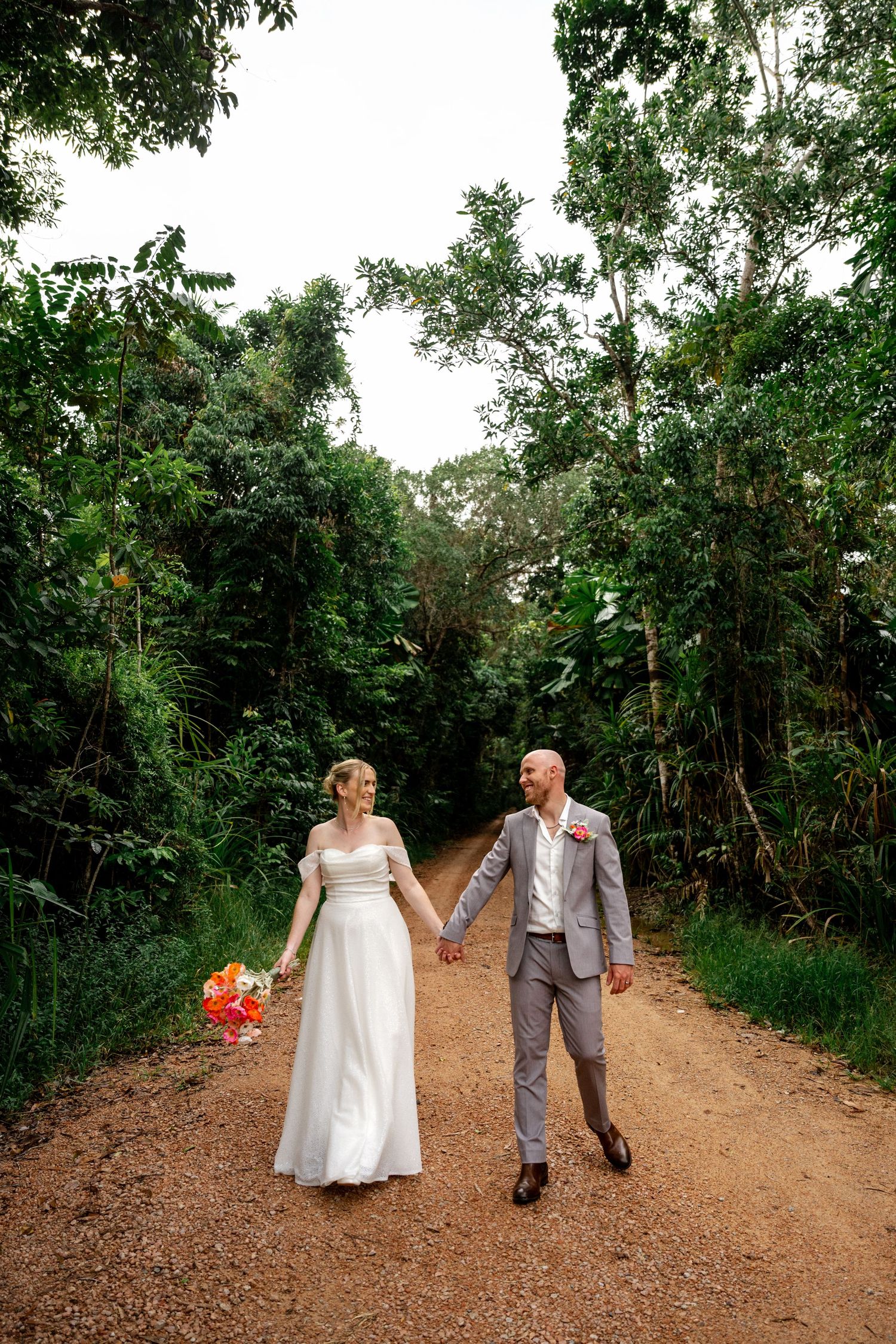 Bride and groom holding hands and gazing at each other on a dirt road surrounded by tropical forest.