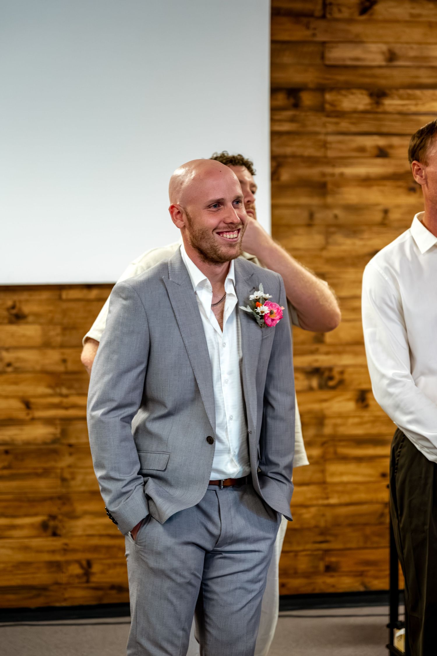 Smiling bald groom in grey suit with pink boutonniere standing at wedding ceremony venue.