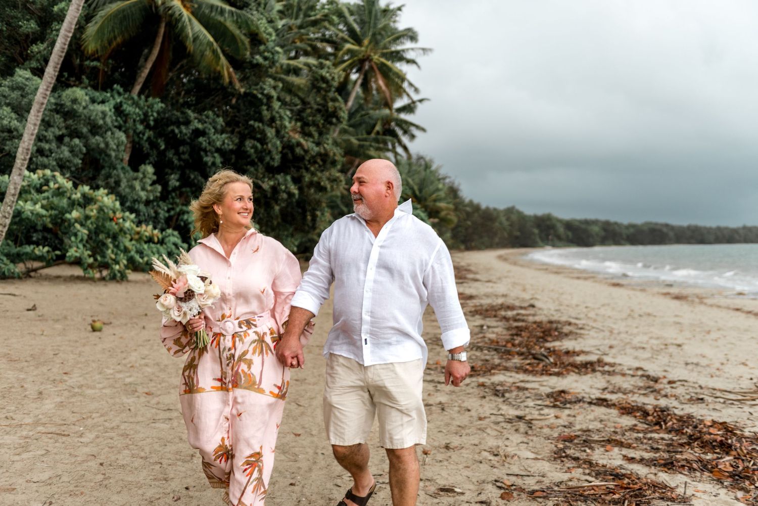 Smiling mature couple holding hands and strolling along a tropical beach with lush palm trees.