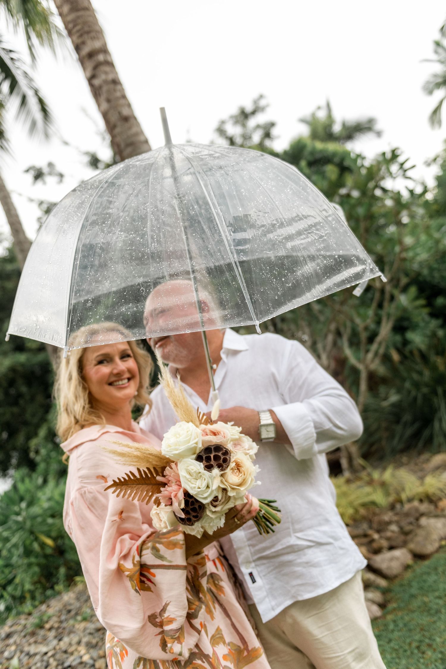 Smiling mature couple under a clear bubble umbrella holding a beautiful floral bouquet outdoors.