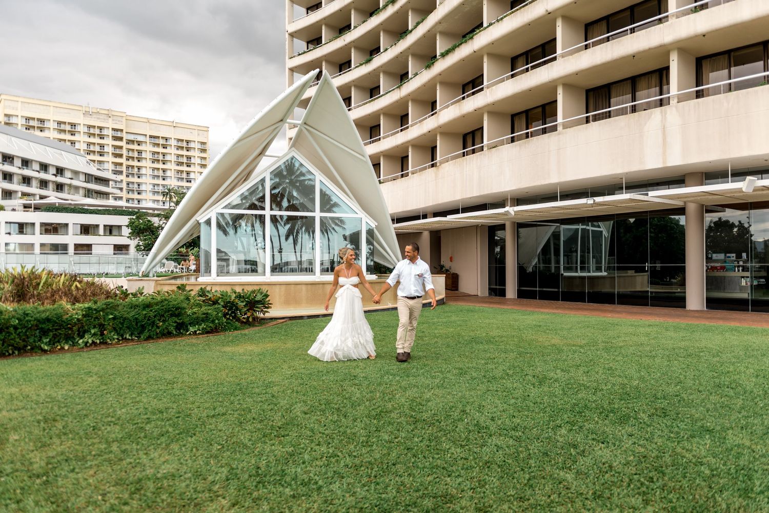 Bride and groom walk hand in hand across green lawn in front of modern hotel with glass chapel.