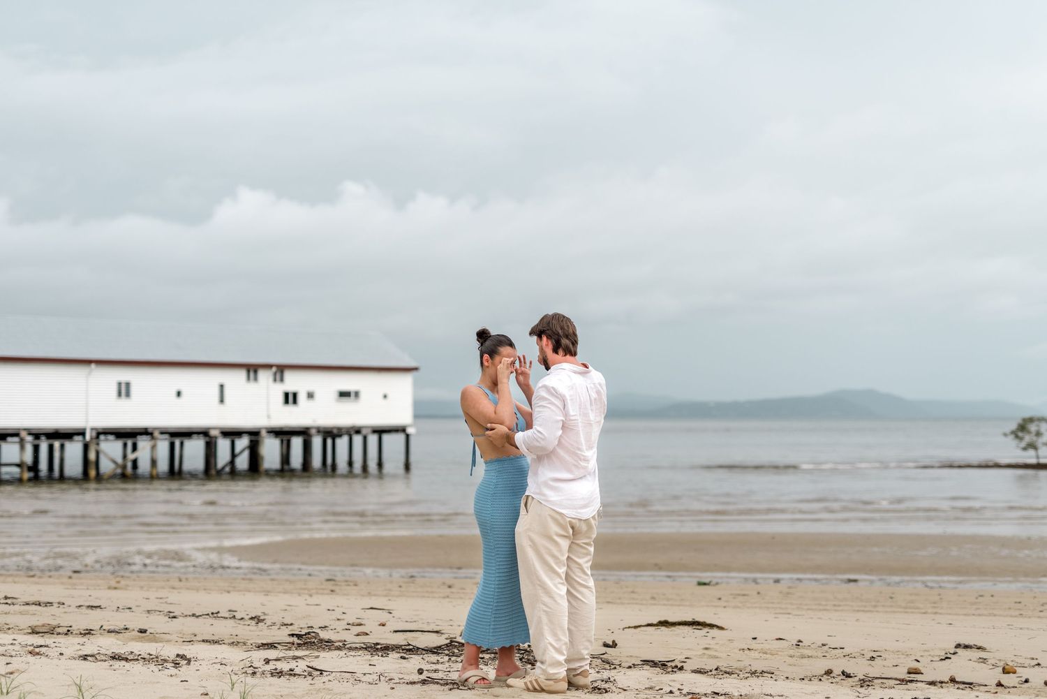 Couple sharing a romantic moment on a sandy beach with a white pier building in the background on an overcast day.