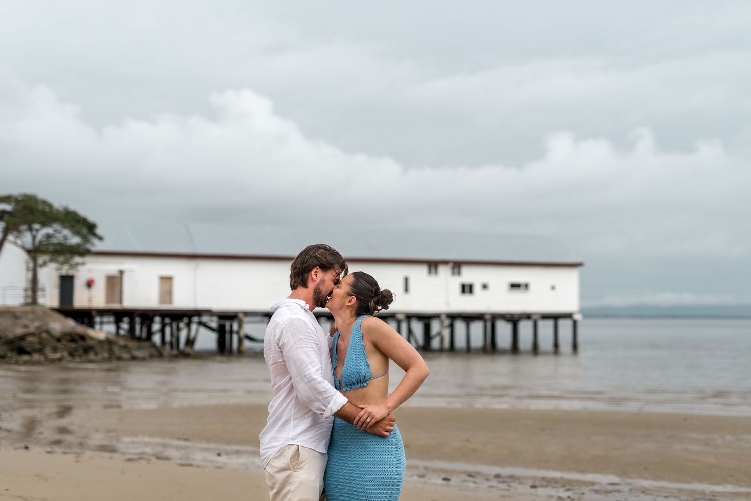 Couple sharing a romantic kiss on a sandy beach with a white building on stilts over the water behind them.