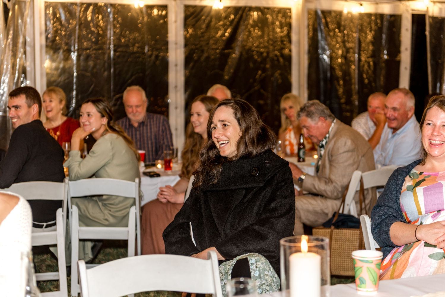 Wedding guests laughing and enjoying themselves at a candlelit reception inside a white draped tent at night.