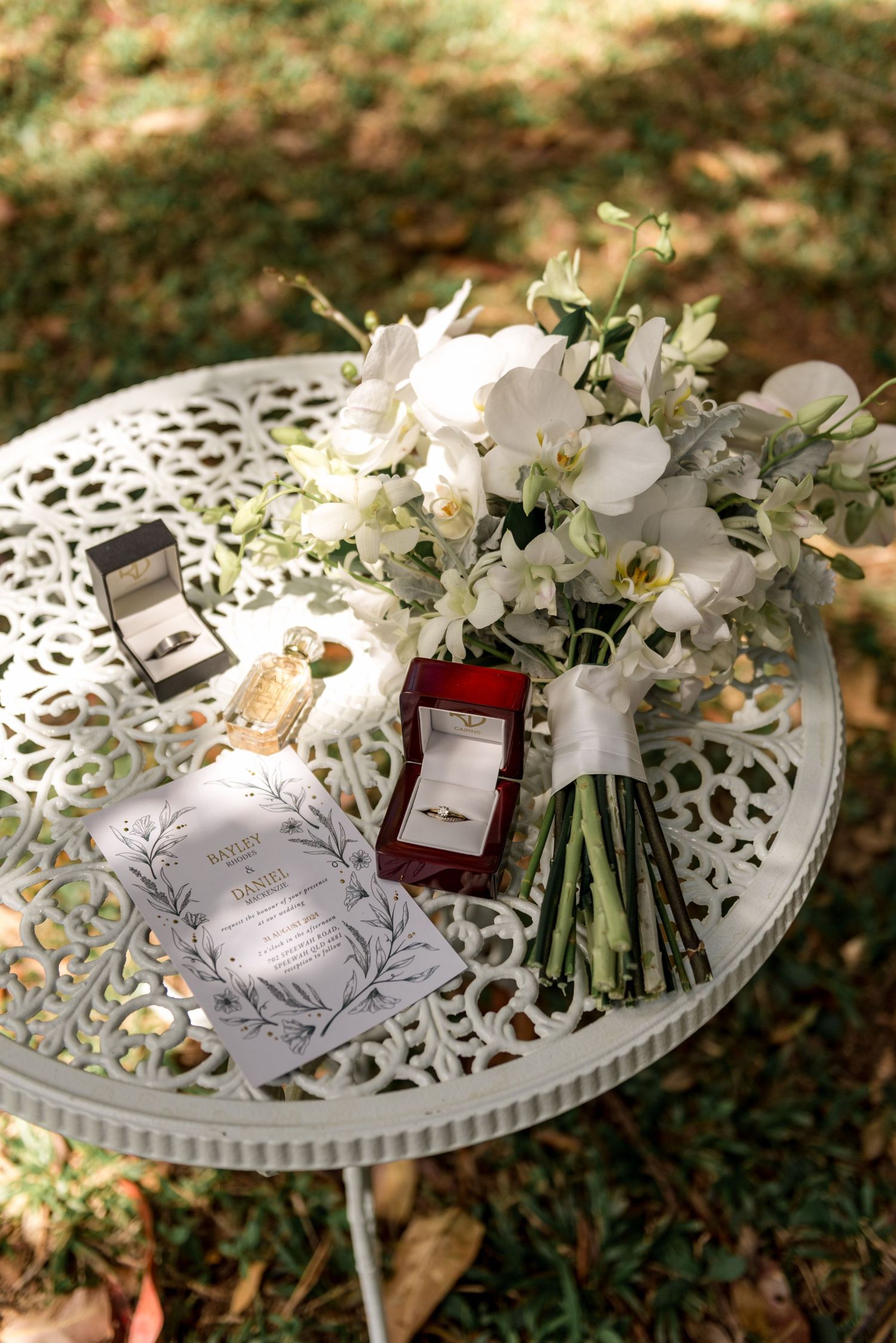 Wedding details styled on ornate white table with orchid bouquet, rings, invitation, and perfume.