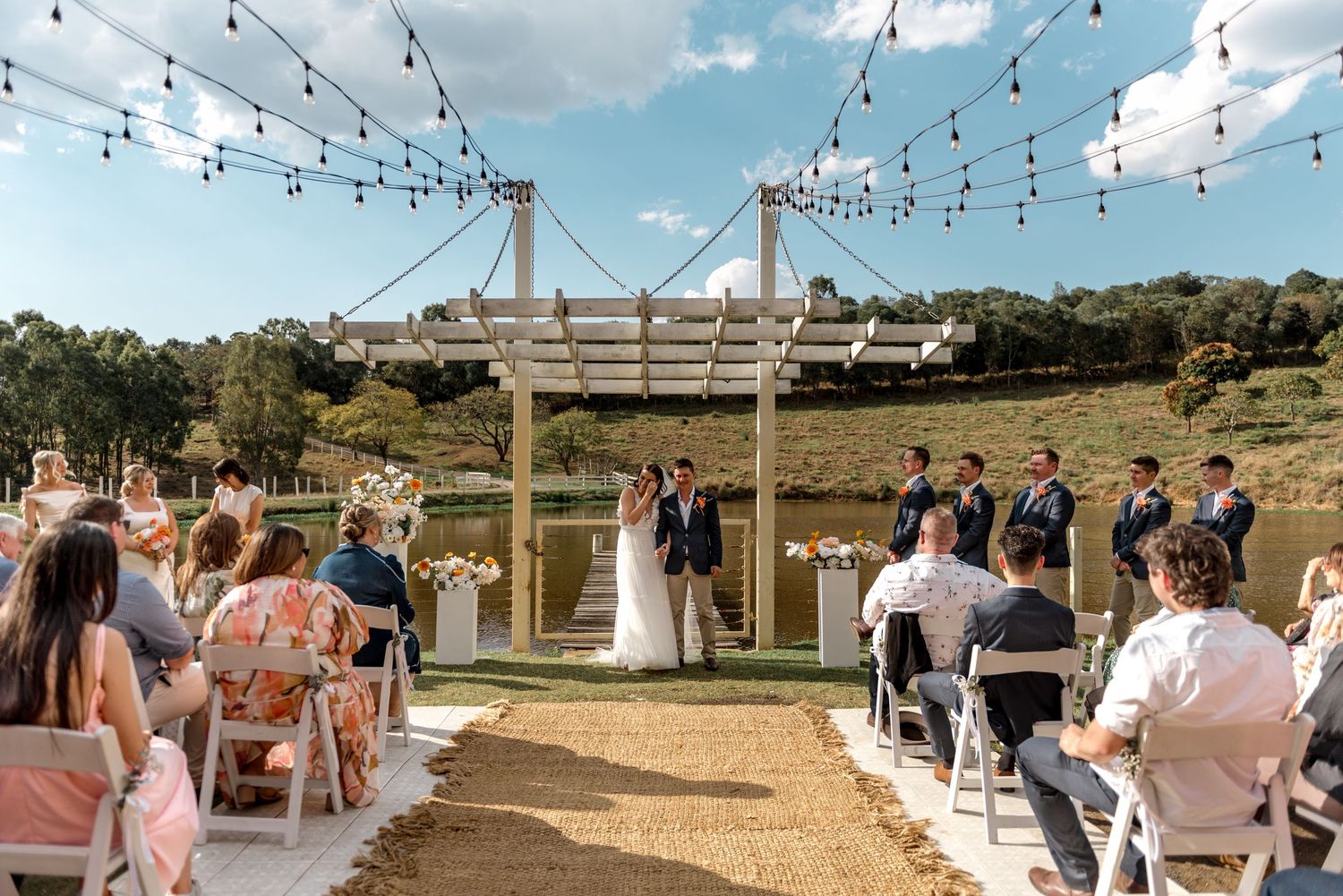 Wedding ceremony at a lakeside venue with string lights, white chairs, and bridal party standing at the altar.