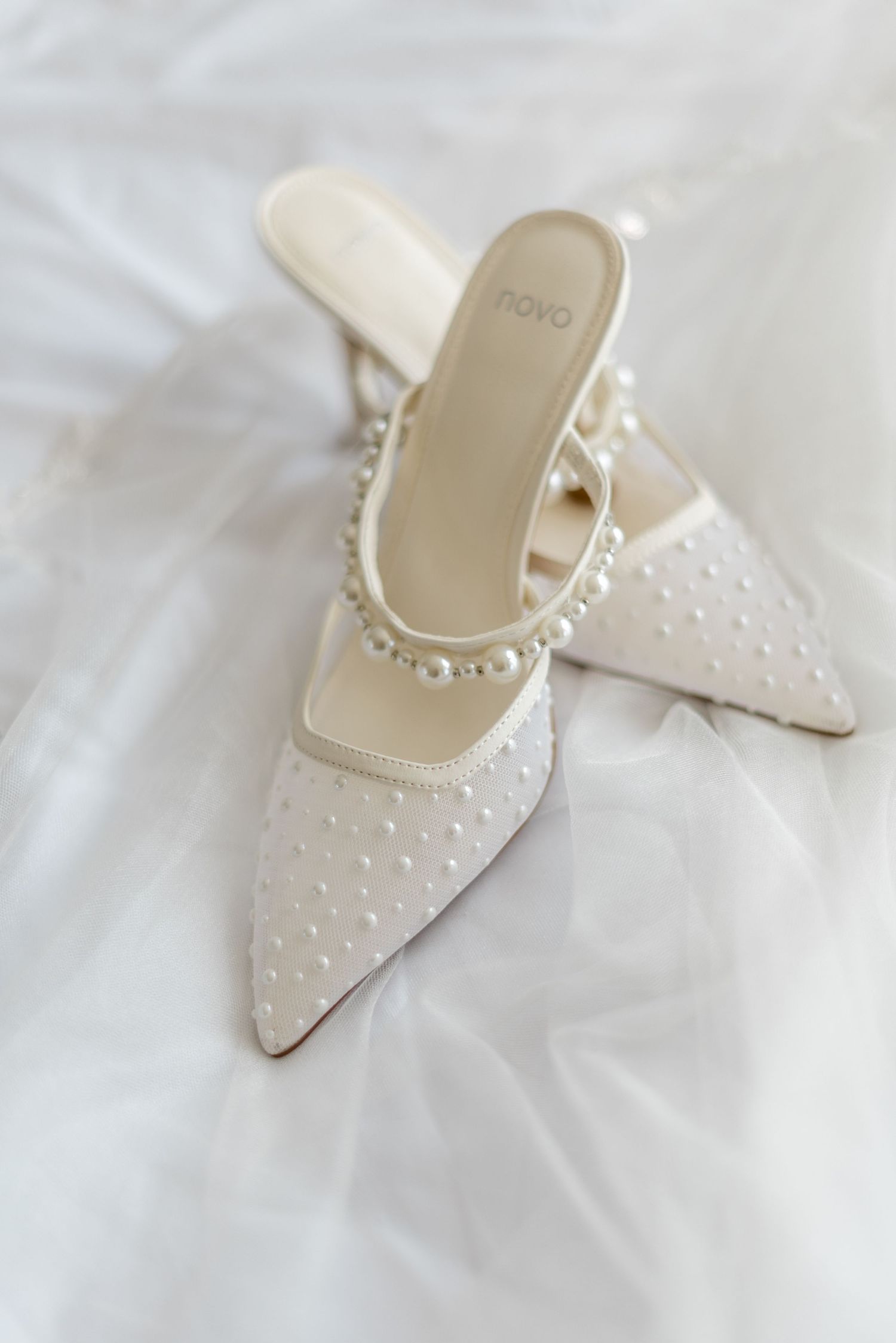 Elegant white pearl-embellished pointed-toe bridal mule heels resting on soft tulle fabric.