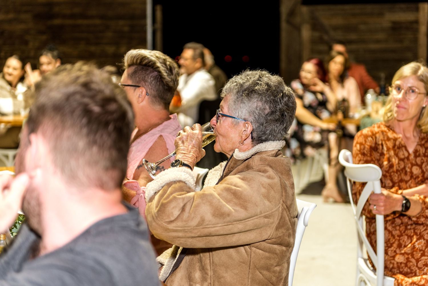 Wedding guest sips wine in rustic barn reception venue surrounded by other seated guests.