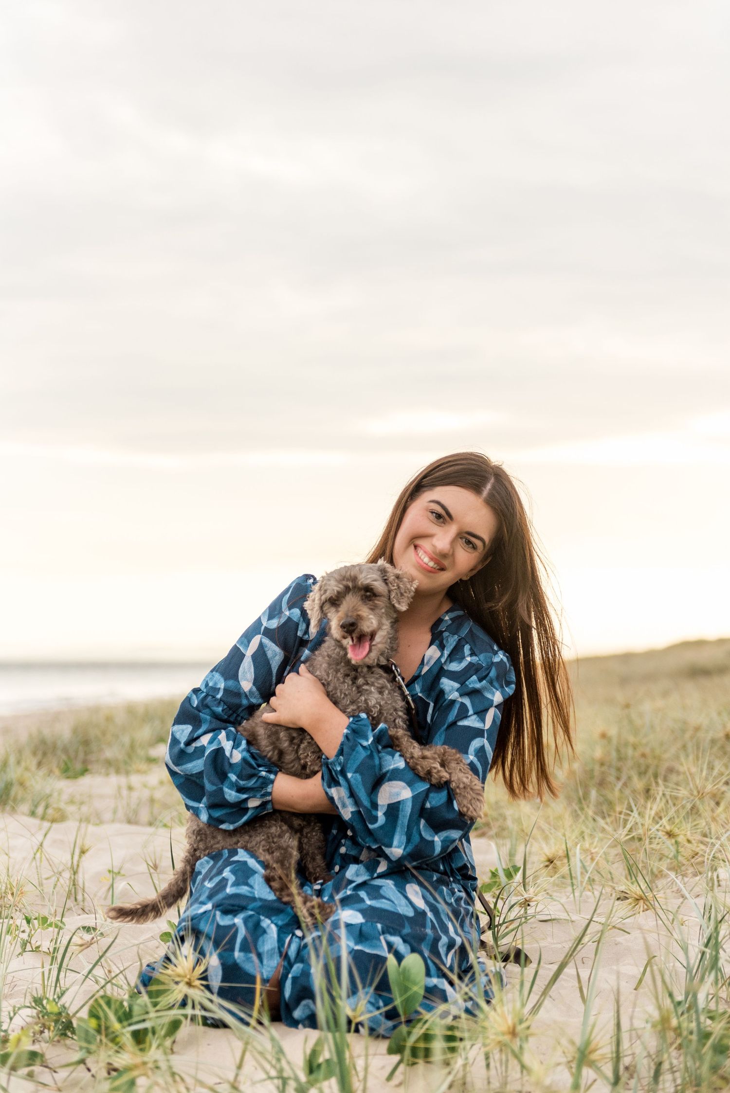 Smiling woman in blue dress cuddling grey fluffy dog on sandy beach dunes.