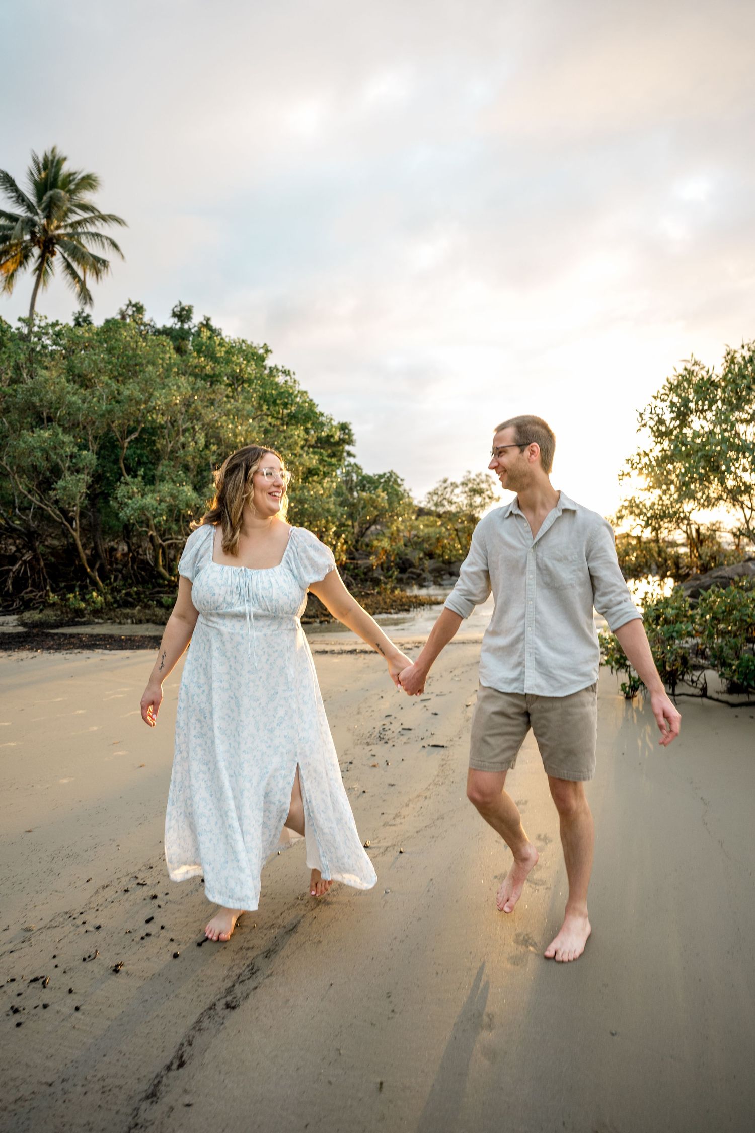 Happy couple holding hands and walking barefoot on a sandy beach at sunset with palm trees.