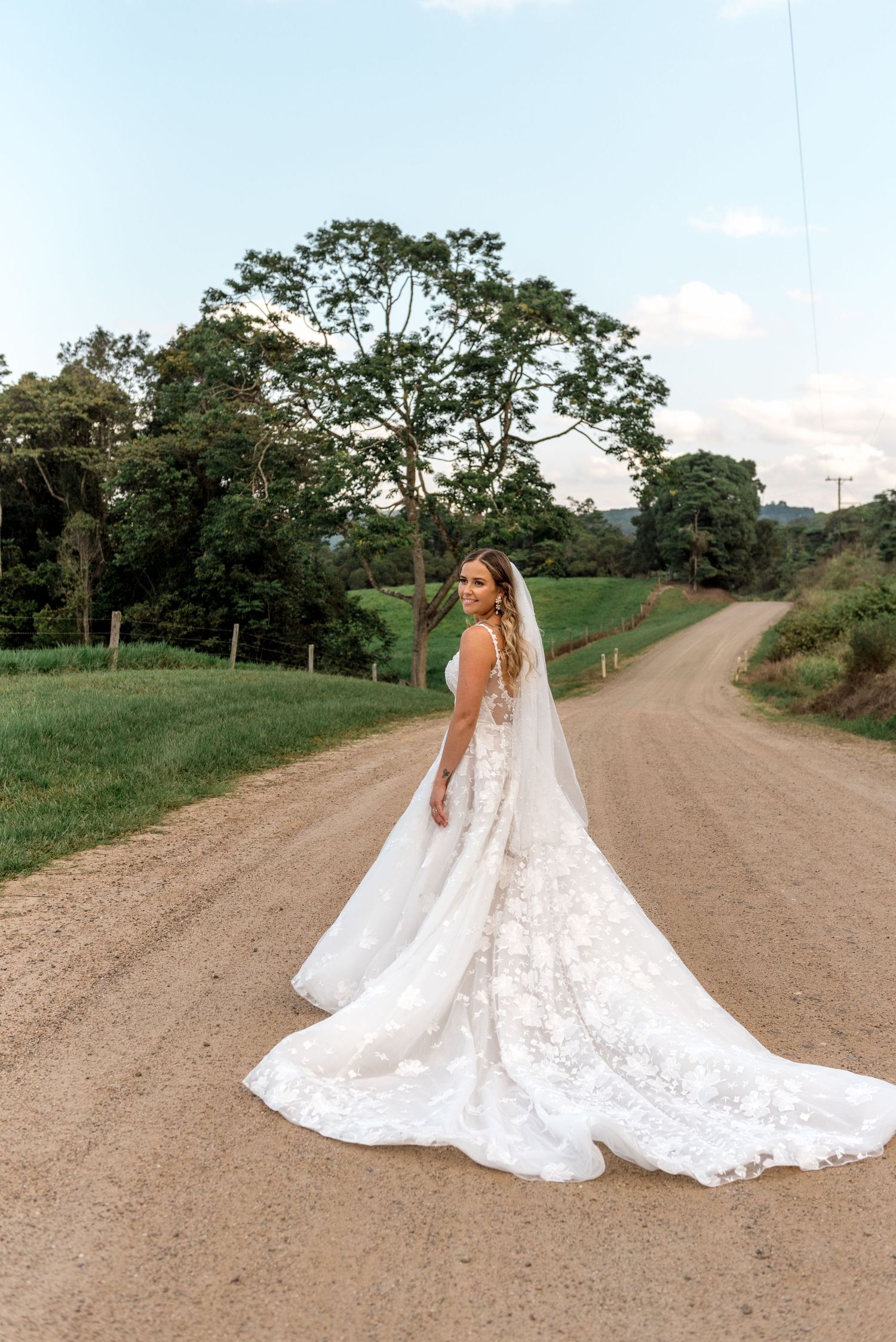 Bride in a floral lace wedding gown with cathedral veil poses on a rustic gravel country road at dusk.