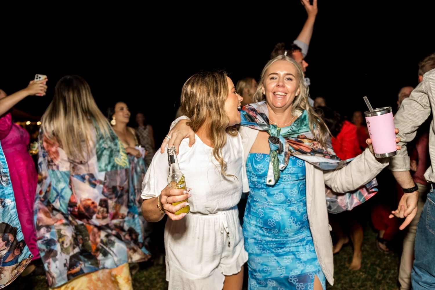 Two women laughing and dancing together at an outdoor night party holding drinks.
