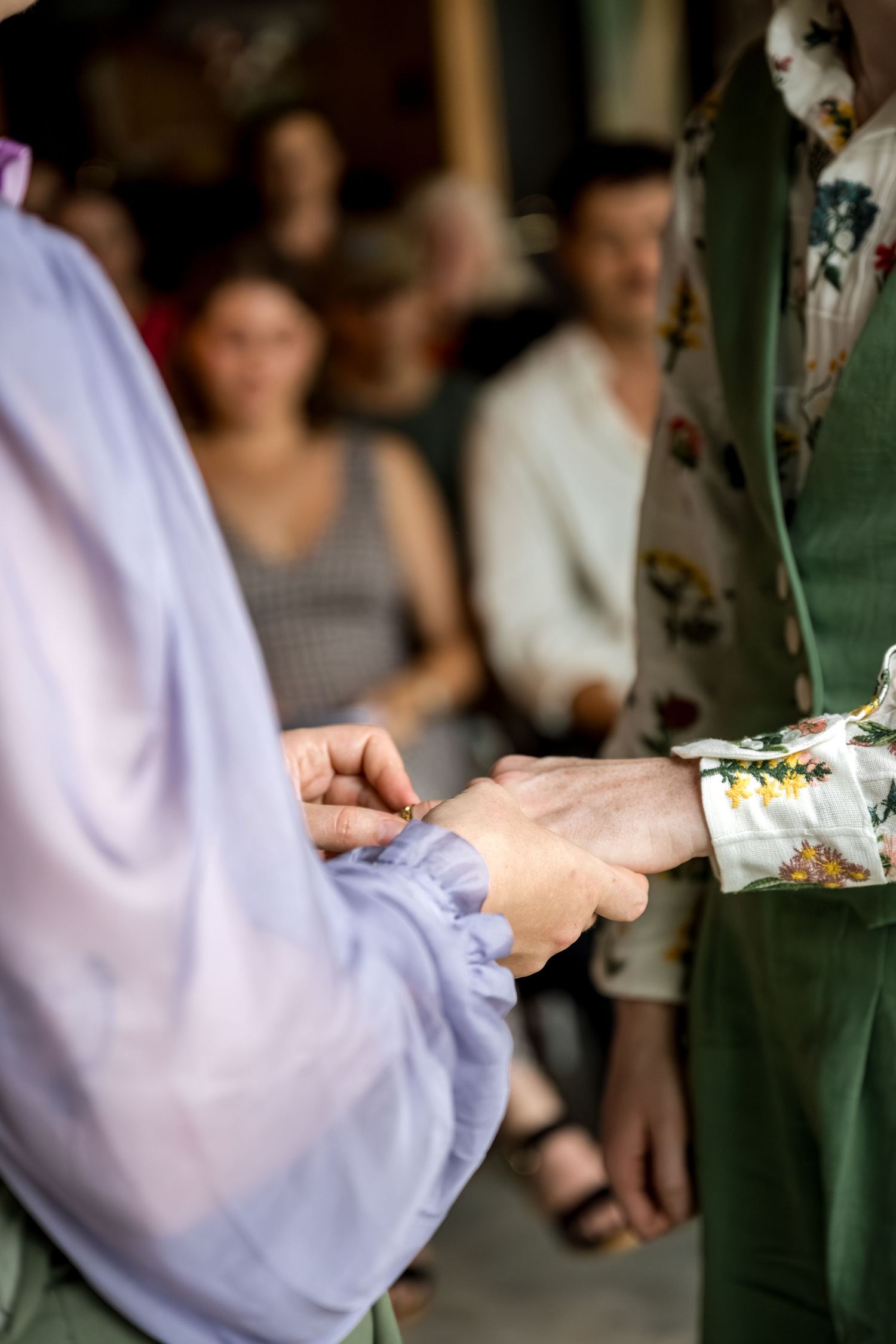 Close-up of two brides holding hands during ring exchange at wedding ceremony with guests in background.