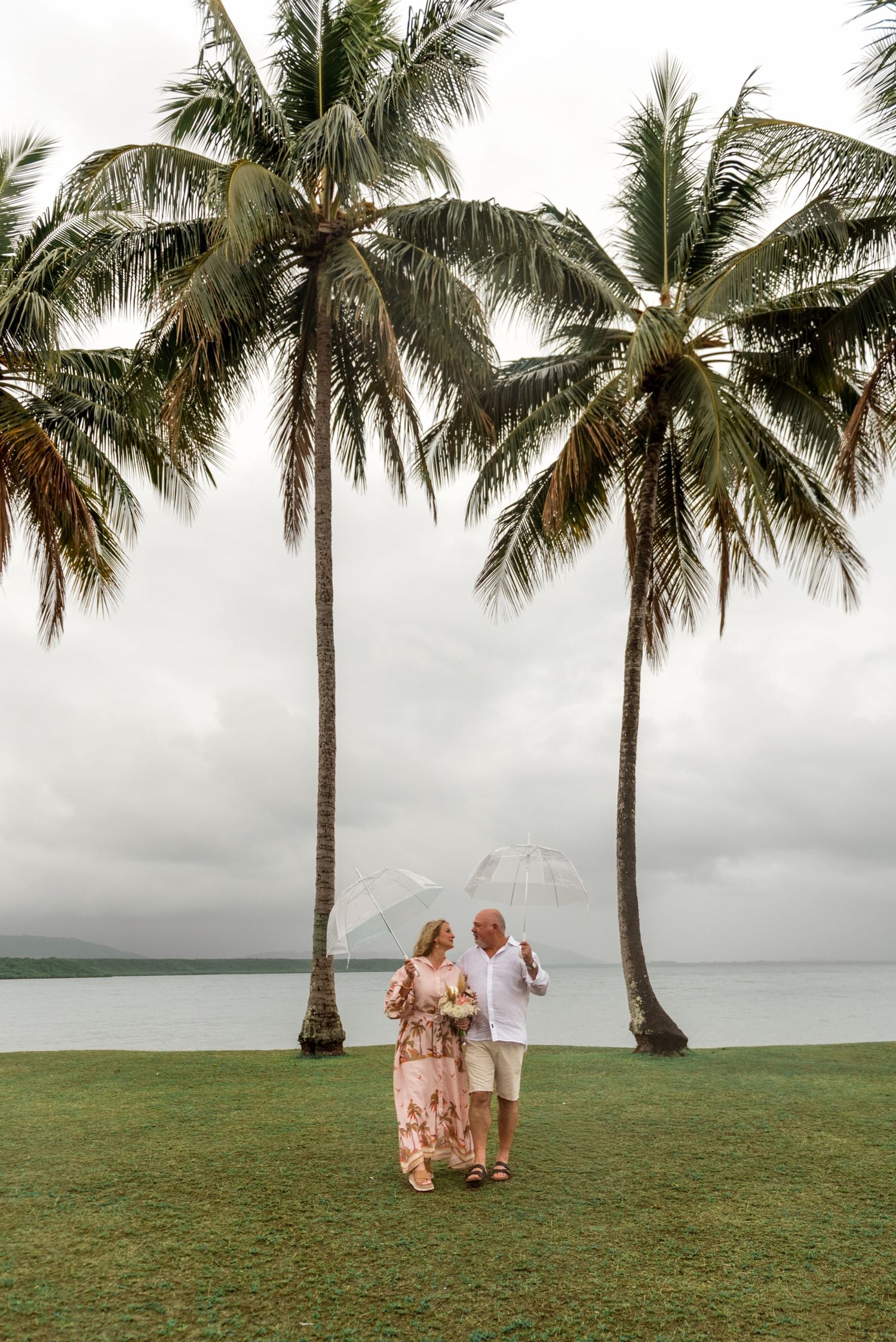 Couple sharing an intimate moment under a clear umbrella between two tall palm trees by the ocean.