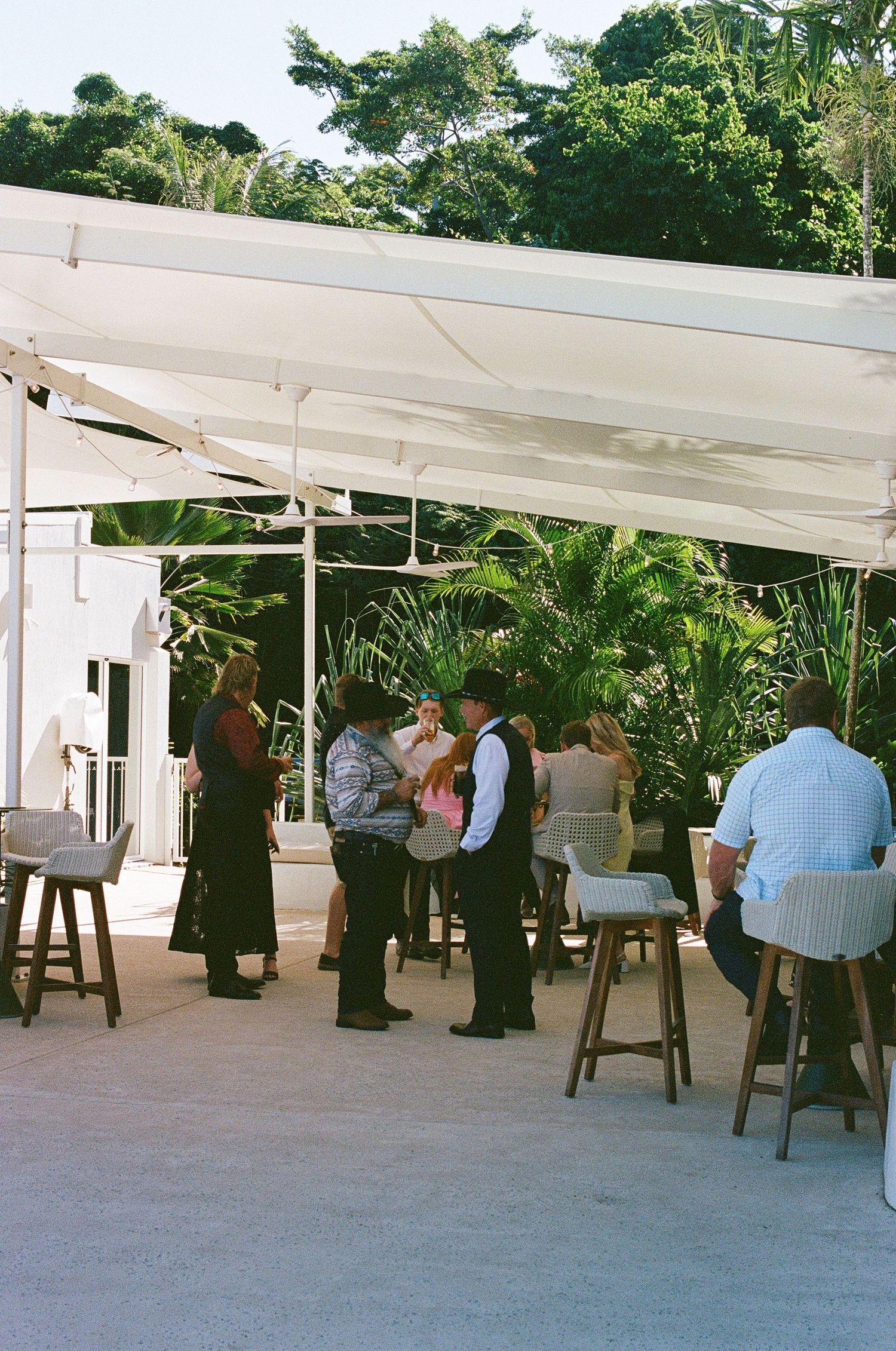 Wedding guests mingling under a white canopy surrounded by lush tropical greenery at an outdoor venue.