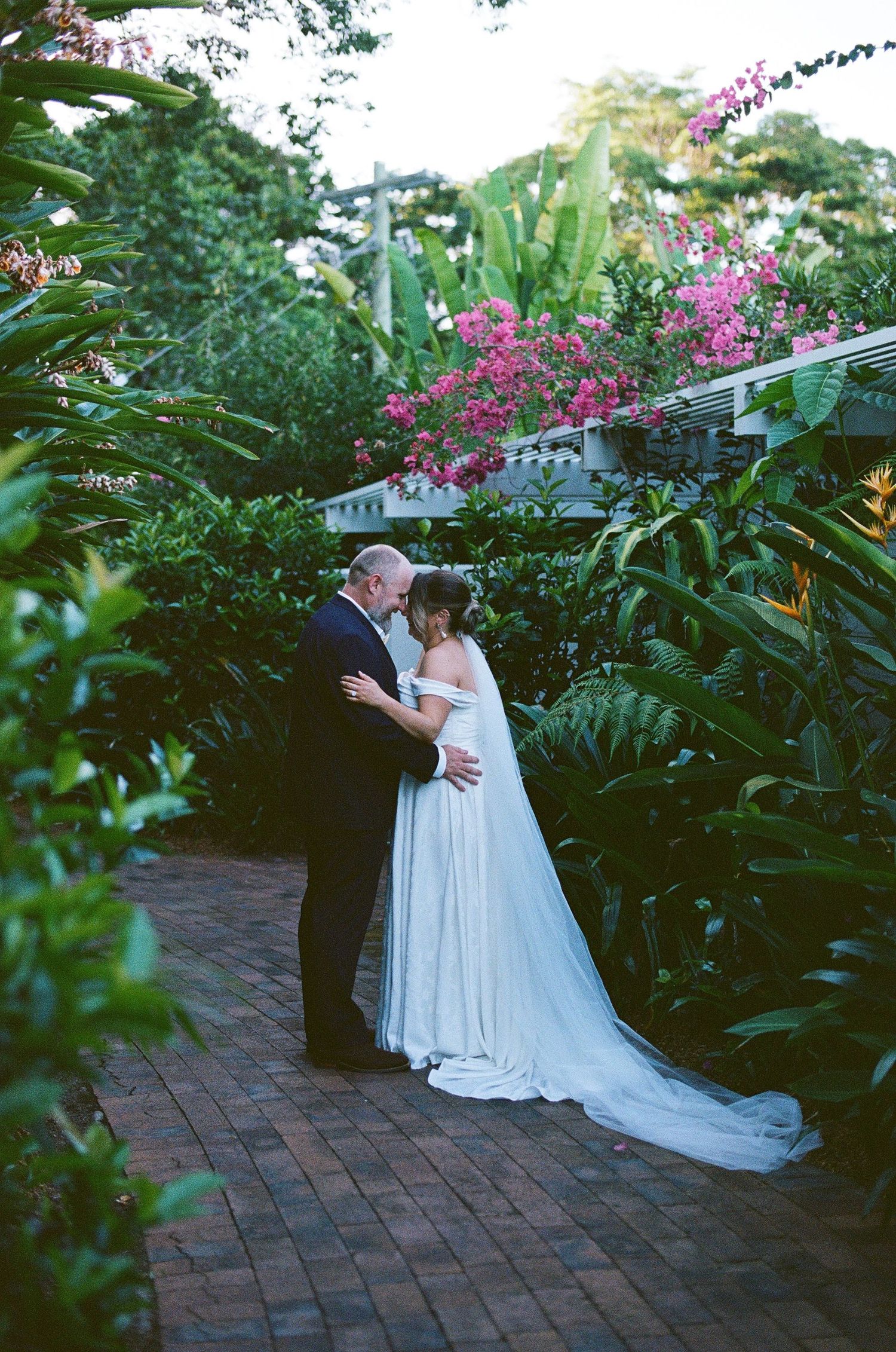 Bride and groom sharing romantic moment on brick pathway surrounded by vibrant tropical gardens and pink flowers.